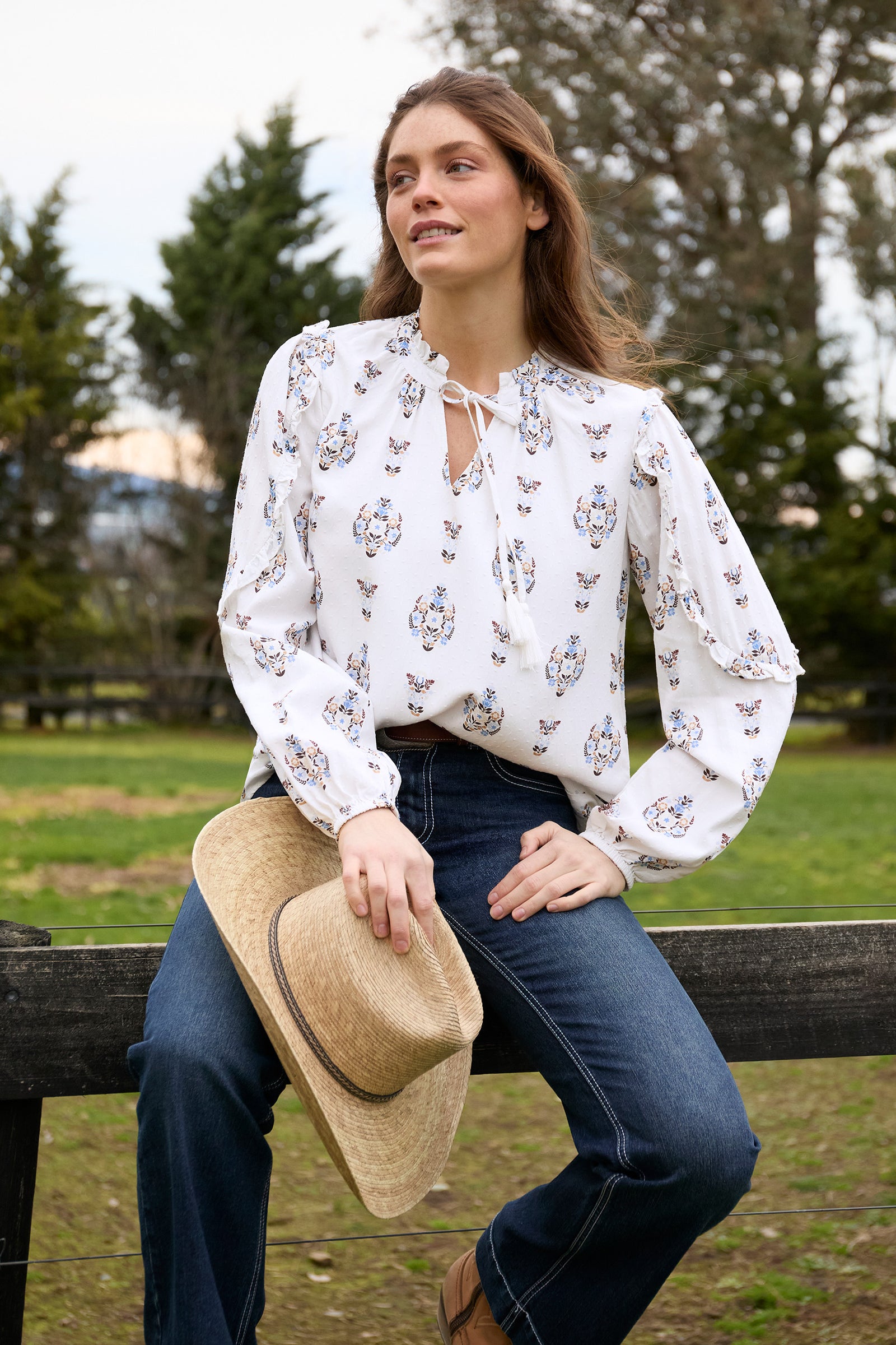 Woman sitting on a wooden bench wearing a white floral blouse and blue jeans, holding a beige hat.