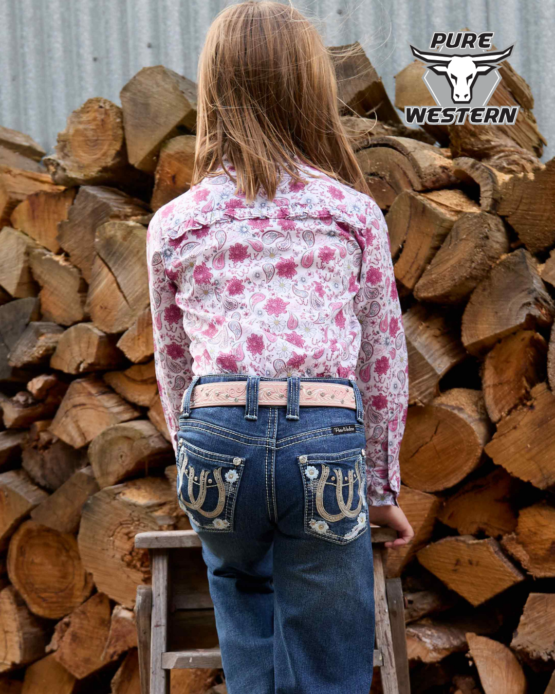 Person wearing a floral shirt and jeans standing in front of stacked logs with 'Pure Western' branding.