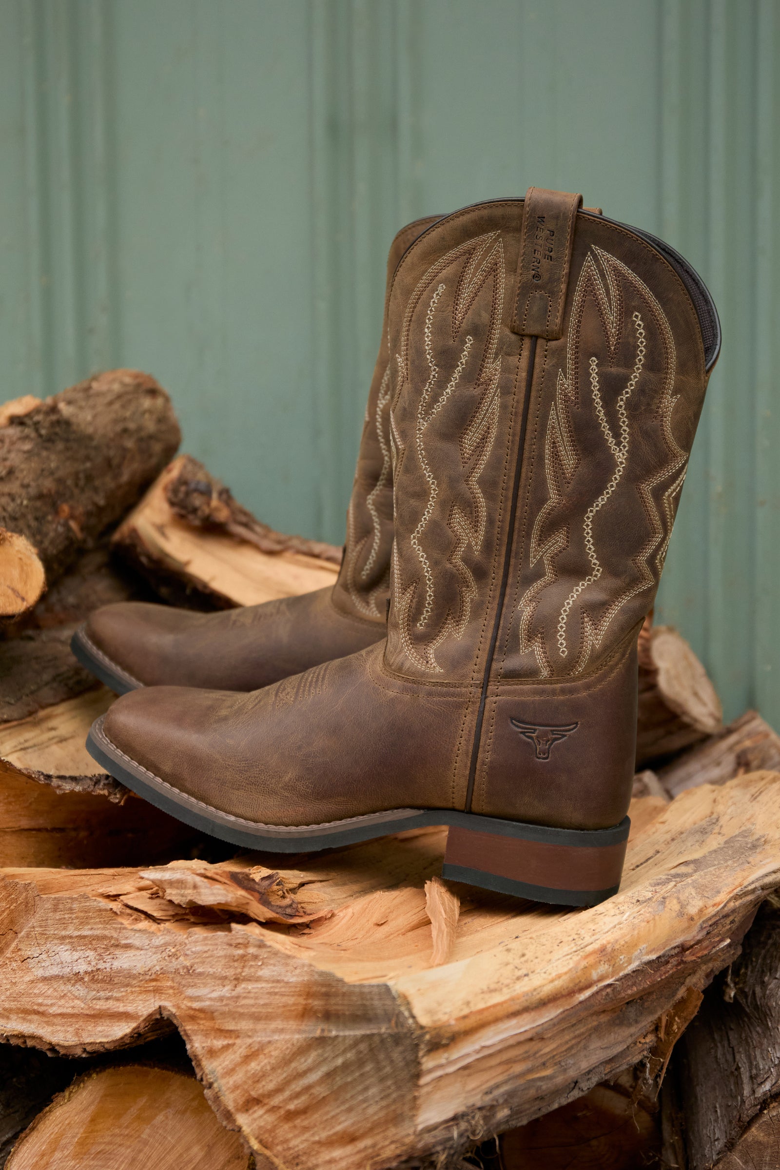 Brown cowboy boots with intricate designs on a wooden log pile against a green metal wall.