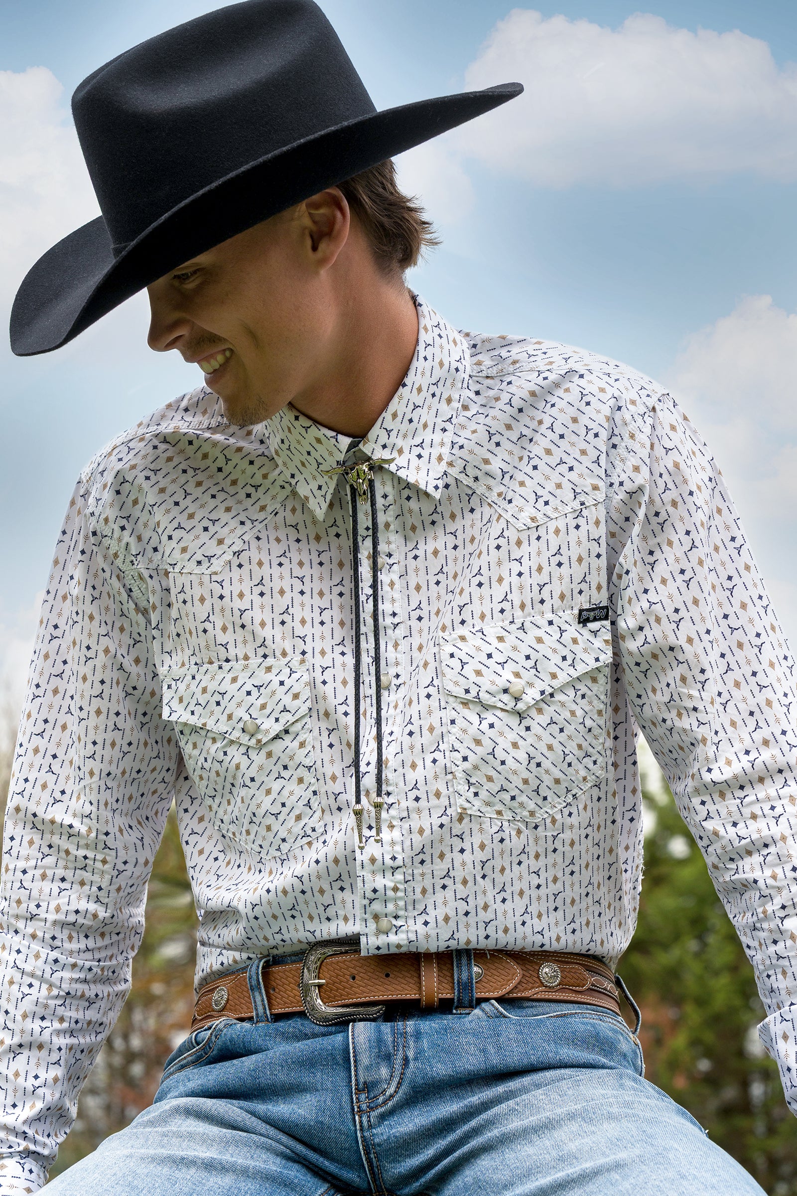 A man wearing a black cowboy hat, a white long-sleeve button-down shirt with a small blue and black geometric pattern, a black bolo tie, and blue jeans, set against a blurred outdoor background of blue sky and greenery.