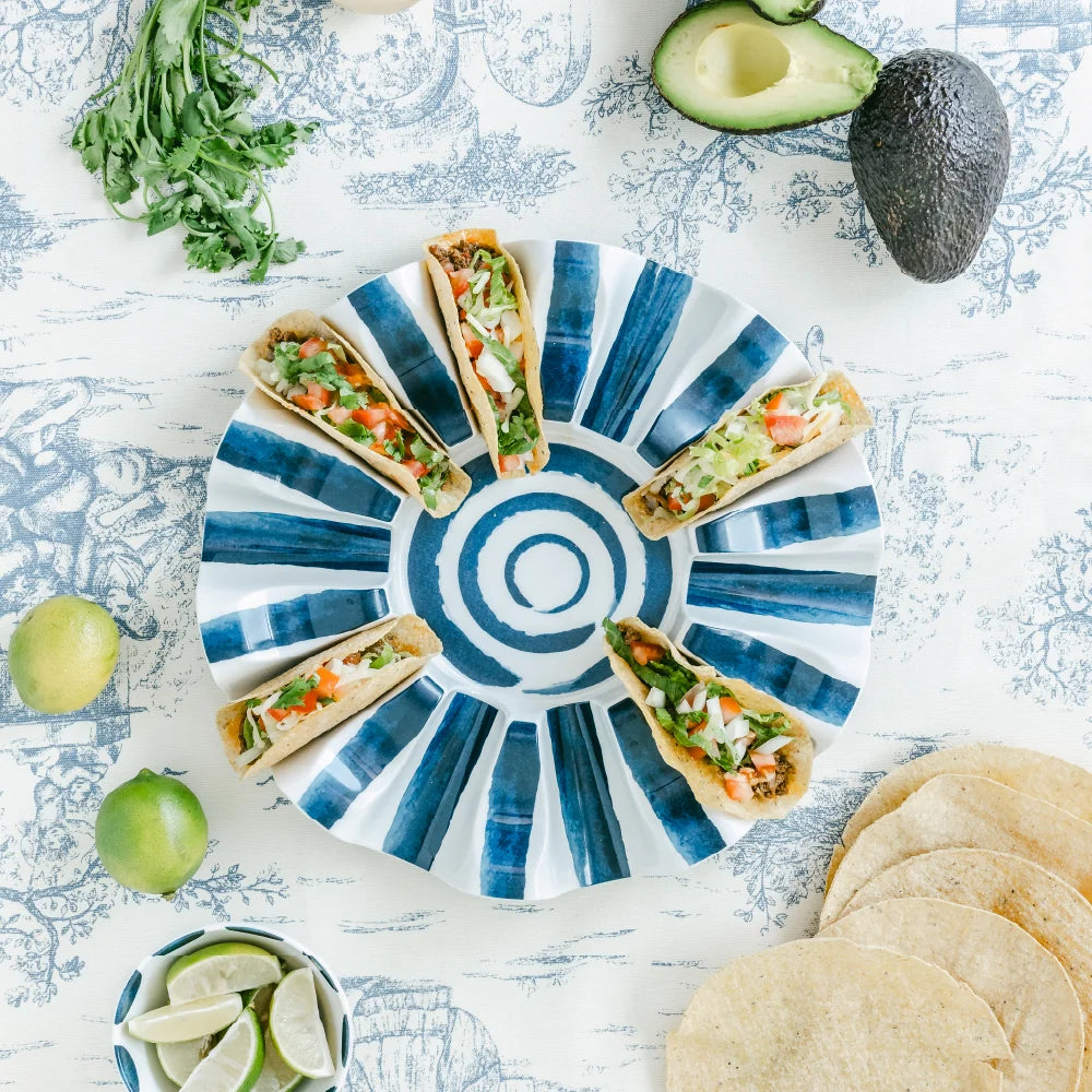 A top-down styled food photo showing five tacos arranged on a blue-and-white striped plate, surrounded by limes, cilantro, avocado, tortilla chips, and a patterned tabletop.