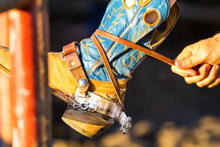 Roughstock Rosin Strapped AF Boot and Spur Ties. Close-up of a cowboy boot with a spur being adjusted, against a blurred background.