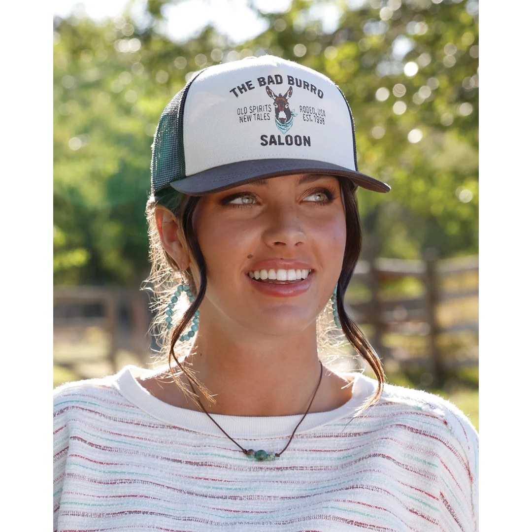 A woman wearing a white and dark green trucker hat with 'THE BAD BURRO SALOON' printed on the front, set against a blurred outdoor background.