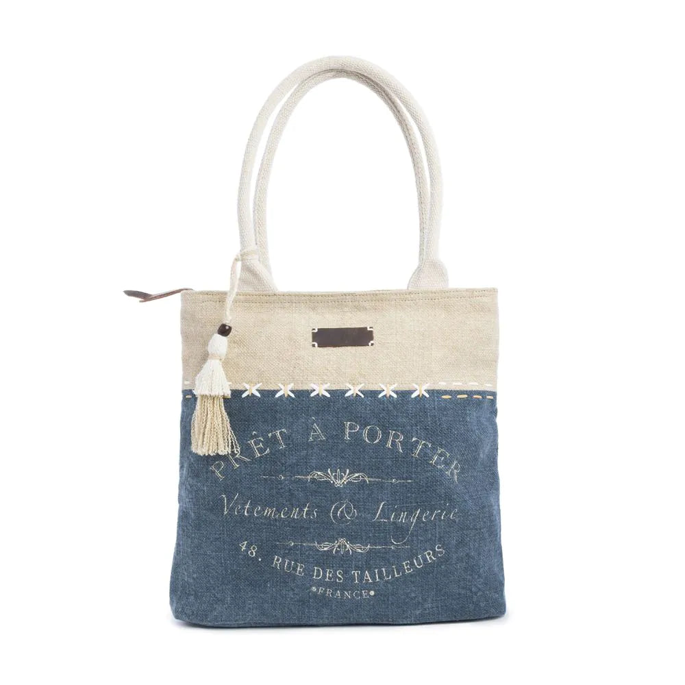 Women's Ports of Call Tote BagA denim blue and wheat colored tote bag with woven canvas handles, featuring decorative stitching and a tassel, with a text design on the front.