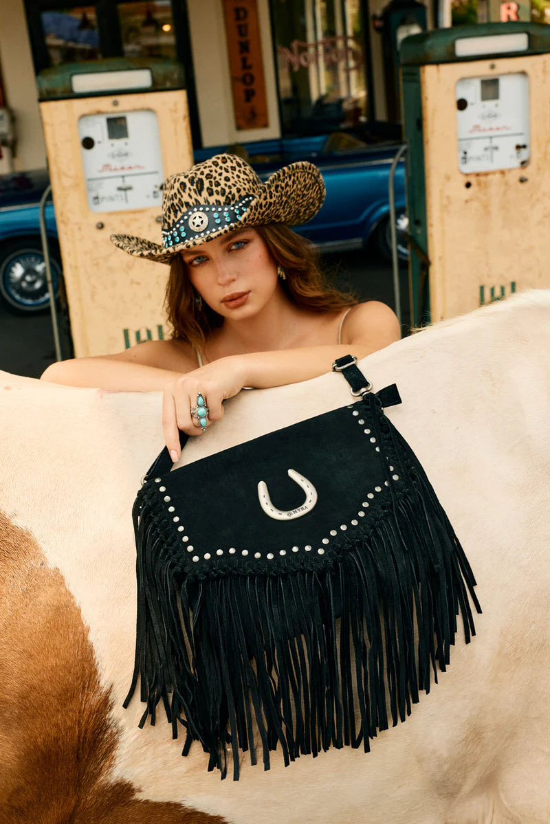 Woman wearing a cowboy hat and holding a black fringe handbag with a horse shoe design, standing next to a vintage car.