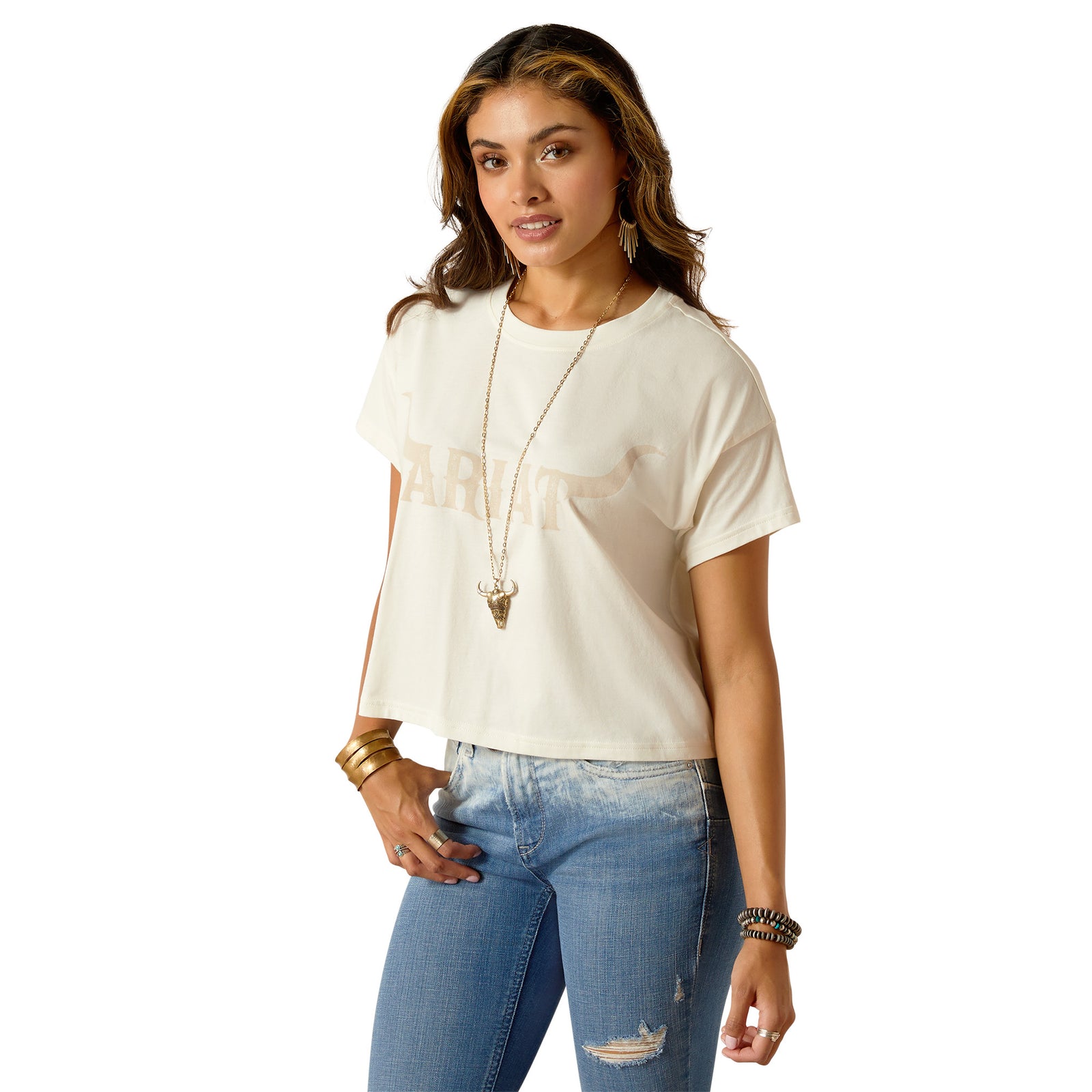 Ariat Women's Wild Hat Cropped S/S T-Shirt. A woman wearing a beige, solid color, cropped T-shirt with a boxy fit, paired with blue denim jeans. She has accessorized with a long necklace and multiple bracelets.