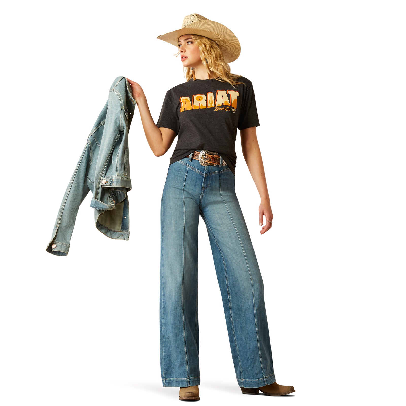 Ariat Women's Postcard T-Shirt. Person wearing a black t-shirt with 'Ariat' text, blue jeans, and a cowboy hat, holding a denim jacket.