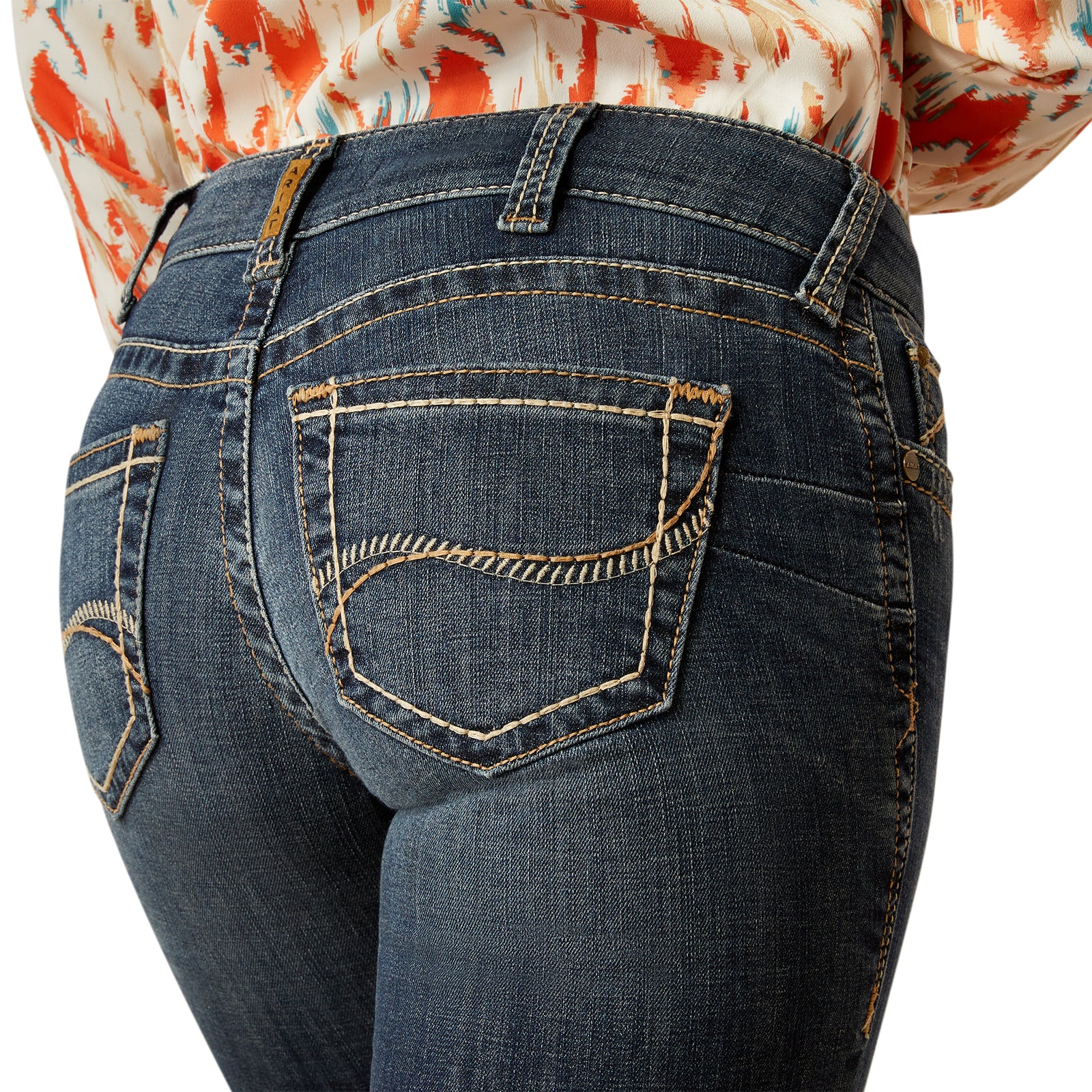 Ariat Women's Lilla Mid Rise Boot Cut Jean. Close-up of blue jeans with a colorful shirt underneath on a white background
