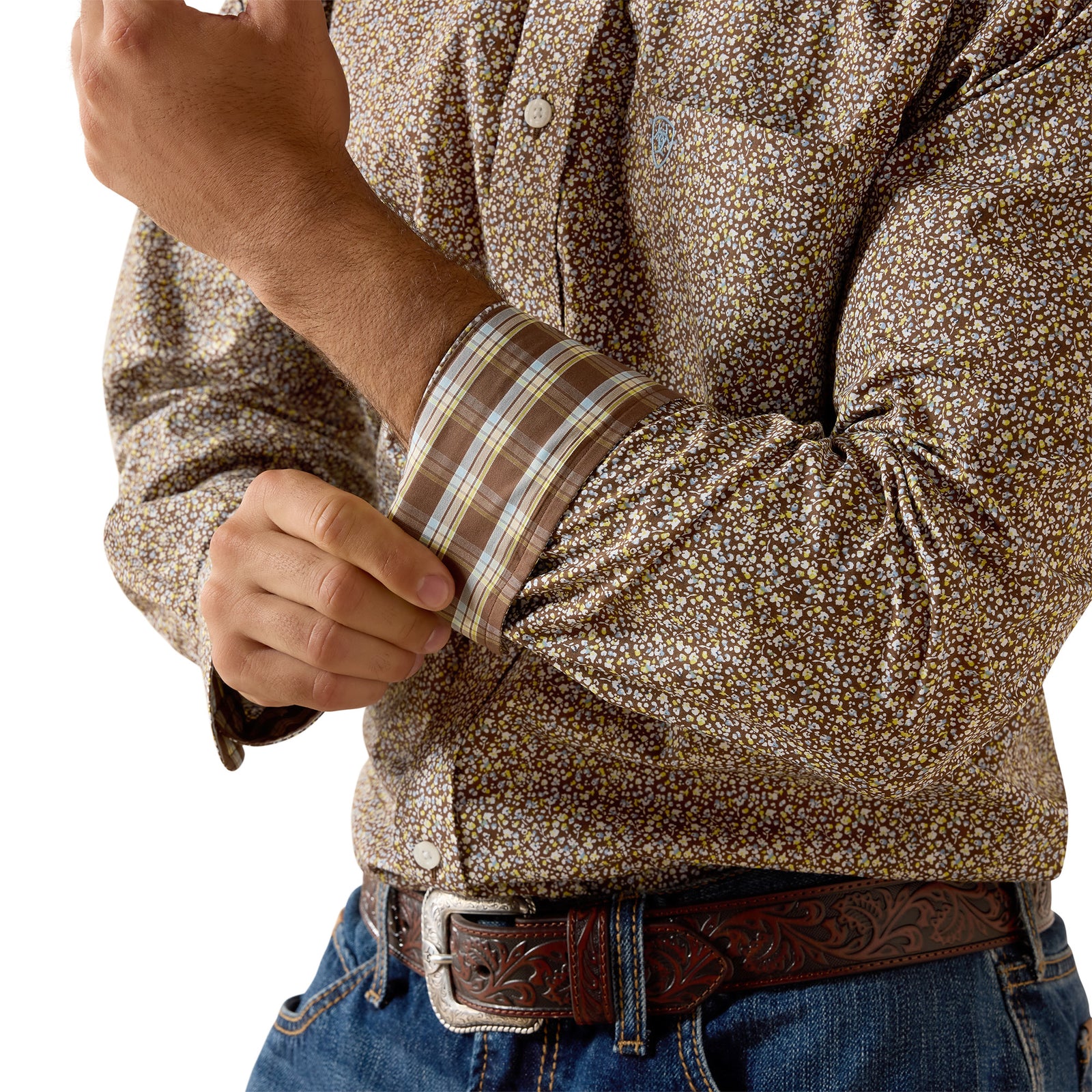 A close-up of a man wearing a brown and beige floral patterned long-sleeve button-down shirt, rolling up his cuff to show the plaid lining, paired with a brown tooled leather belt and blue jeans.