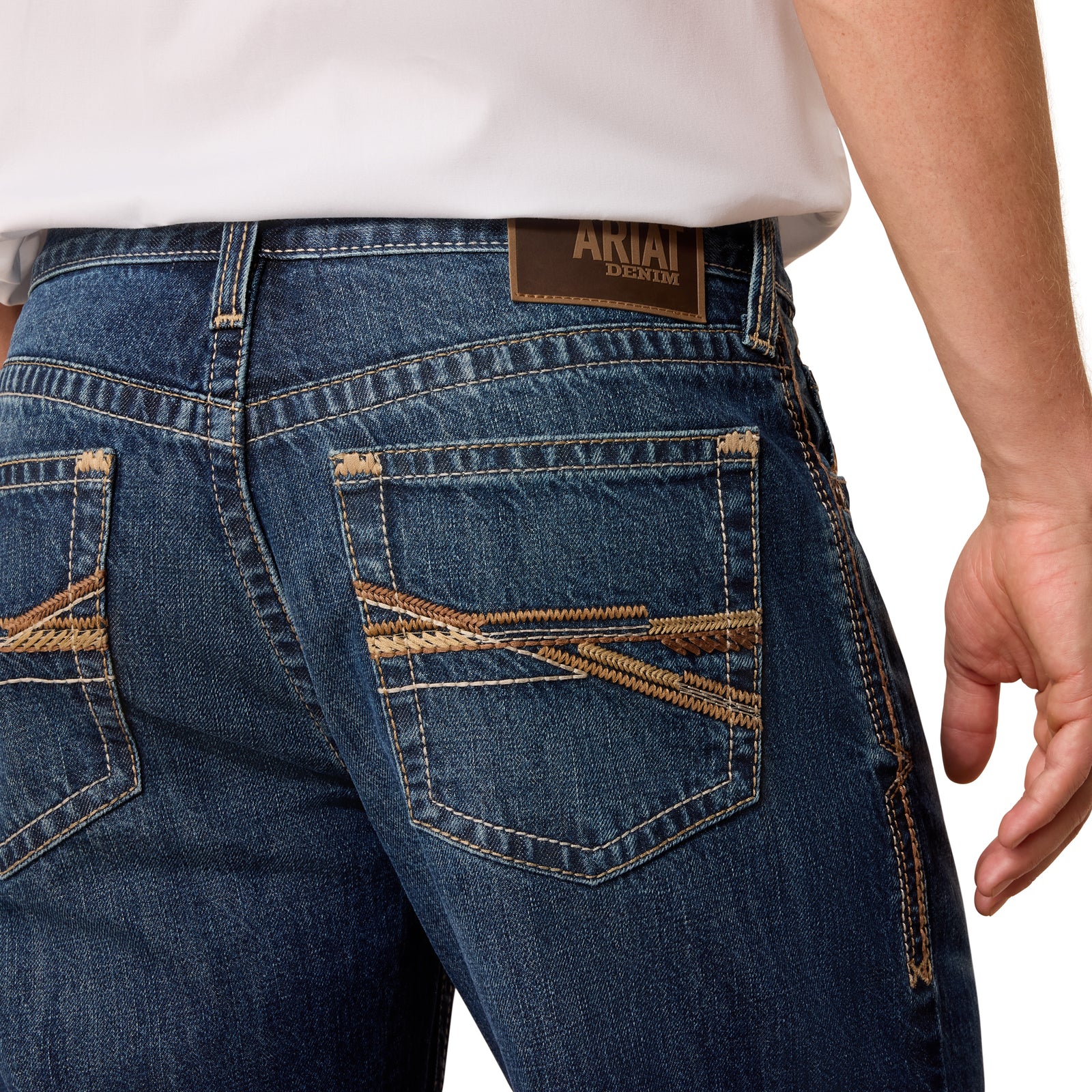Close-up back view of a person wearing blue denim jeans with decorative tan contrast stitching and a brown leather ARIAT DENIM brand patch at the waistband, paired with a white shirt against a light gray background.