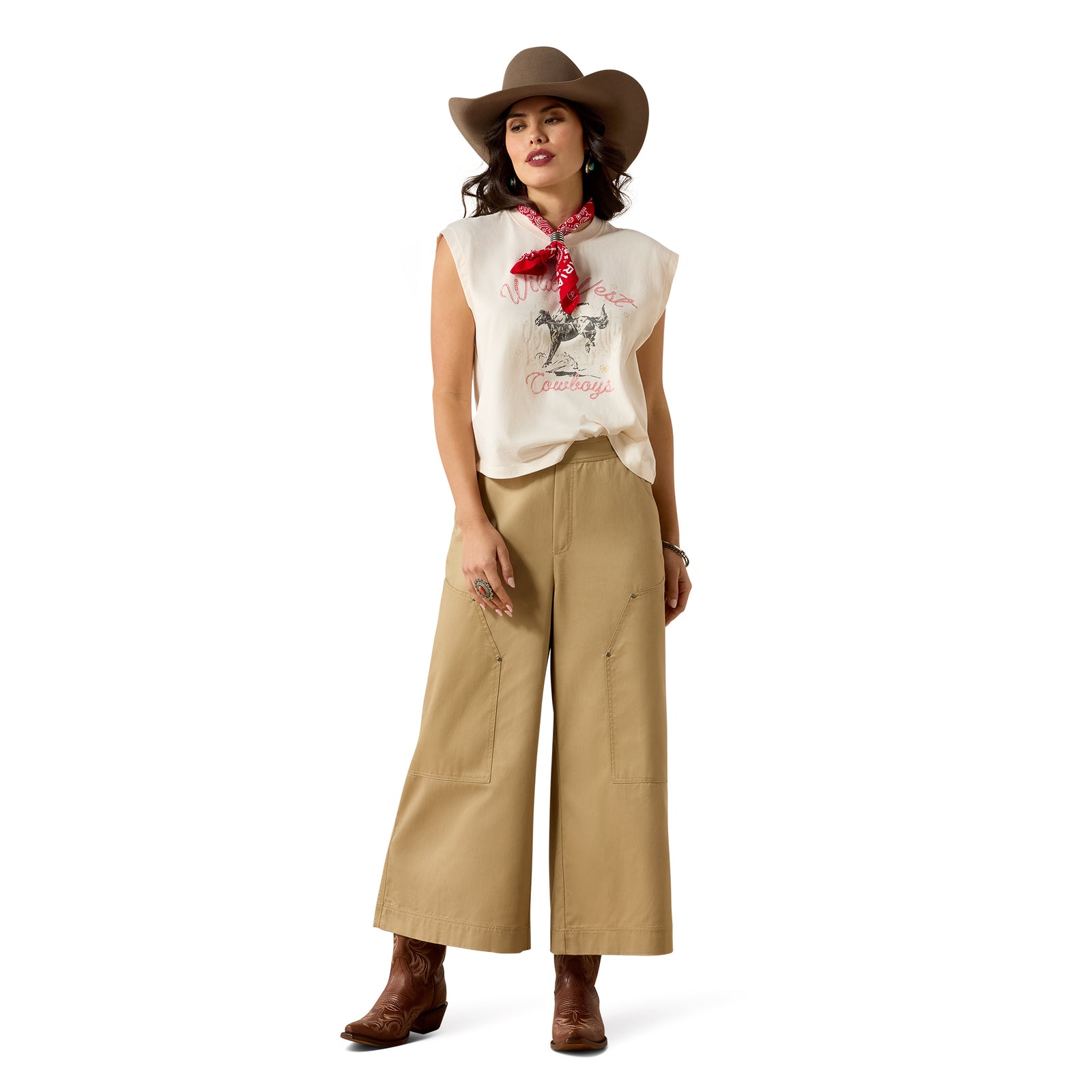 A woman modeling a western-style outfit featuring a cream sleeveless top with a graphic print, beige wide-leg pants, a brown cowboy hat, a red bandana, and brown boots, set against a plain white background.