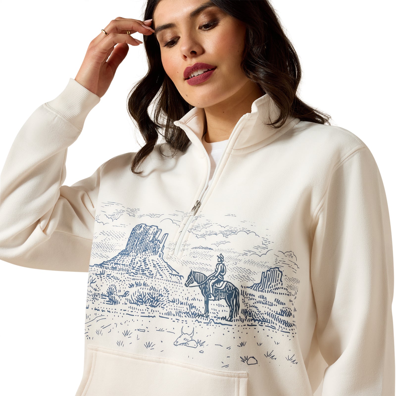 A woman wearing a cream-colored half-zip sweatshirt with a blue western landscape graphic on the front, featuring a rider on a horse and desert mountains.