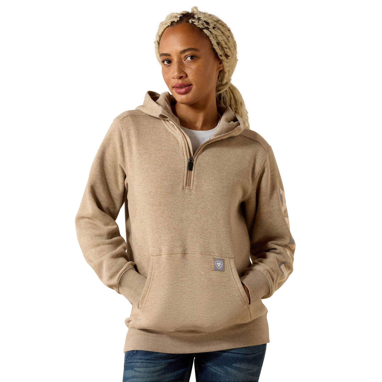 A woman wearing a light tan heathered pullover hoodie with a half-zip neckline, kangaroo pocket, and a small square logo patch near the lower front, paired with blue jeans against a plain white background.