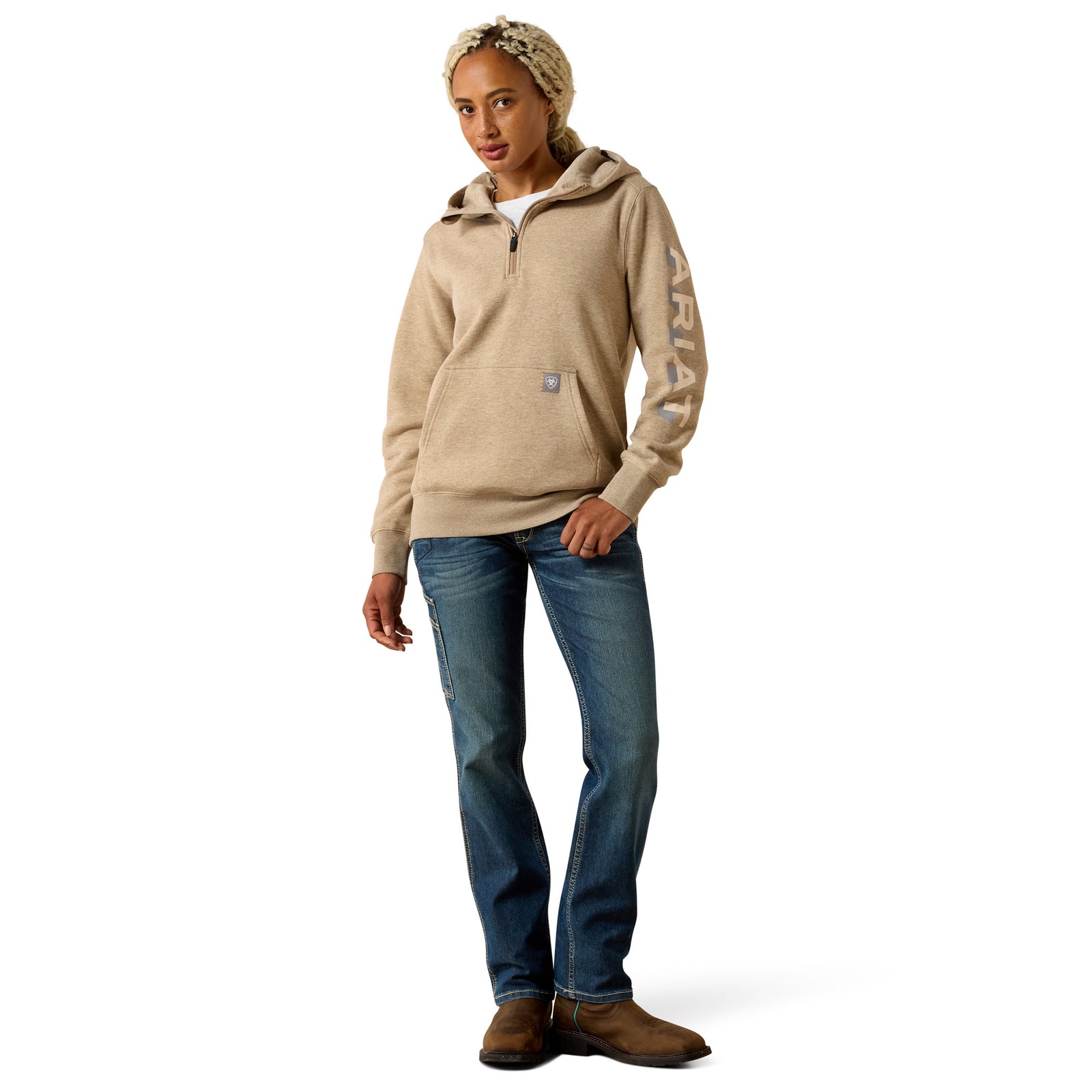 A woman modeling a beige Carhartt quarter-zip hoodie with a vertical logo on the sleeve, paired with blue jeans and brown boots against a white background.