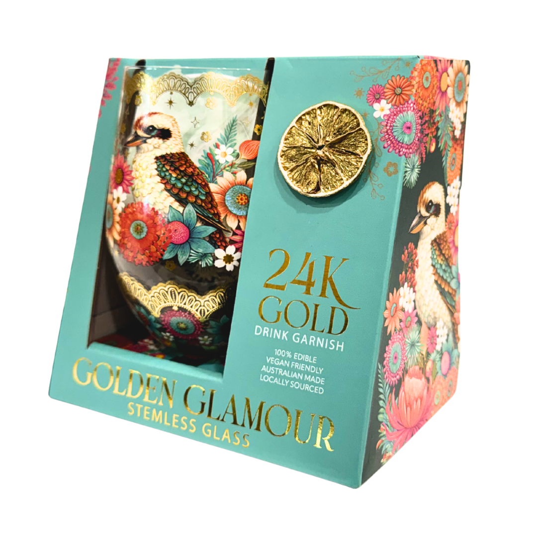 A boxed stemless glass drink garnish set labeled '24K GOLD DRINK GARNISH' and 'GOLDEN GLAMOUR.' The turquoise retail box shows a decorative glass with a kookaburra and floral print visible through a window, plus a dried lemon slice decoration and multicolored floral artwork on the package.