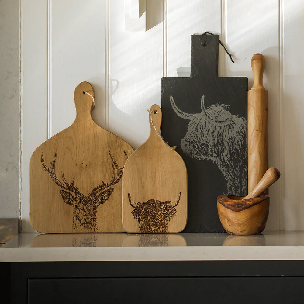 Three wooden cutting boards with engraved animal designs (deer, cow, and Highland cow) are displayed on a kitchen counter alongside a dark slate board, a wooden rolling pin, and a wooden mortar and pestle against a white paneled wall.