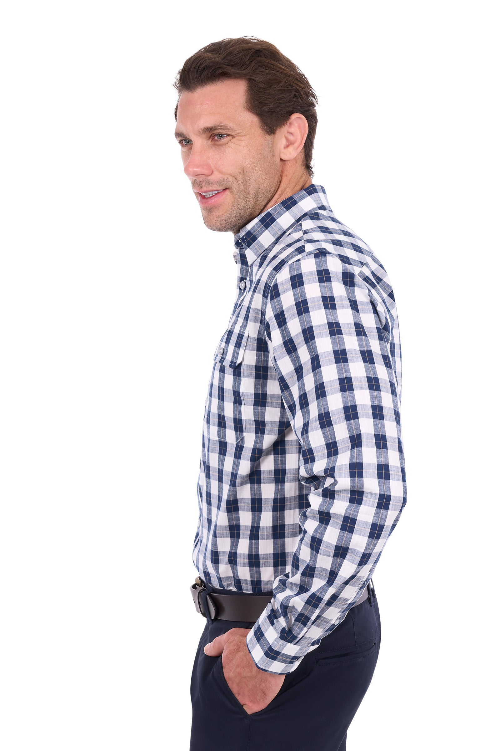 A man in side profile wearing a blue and white checkered long-sleeve button-down shirt, dark trousers, and a black belt, photographed against a solid white background.