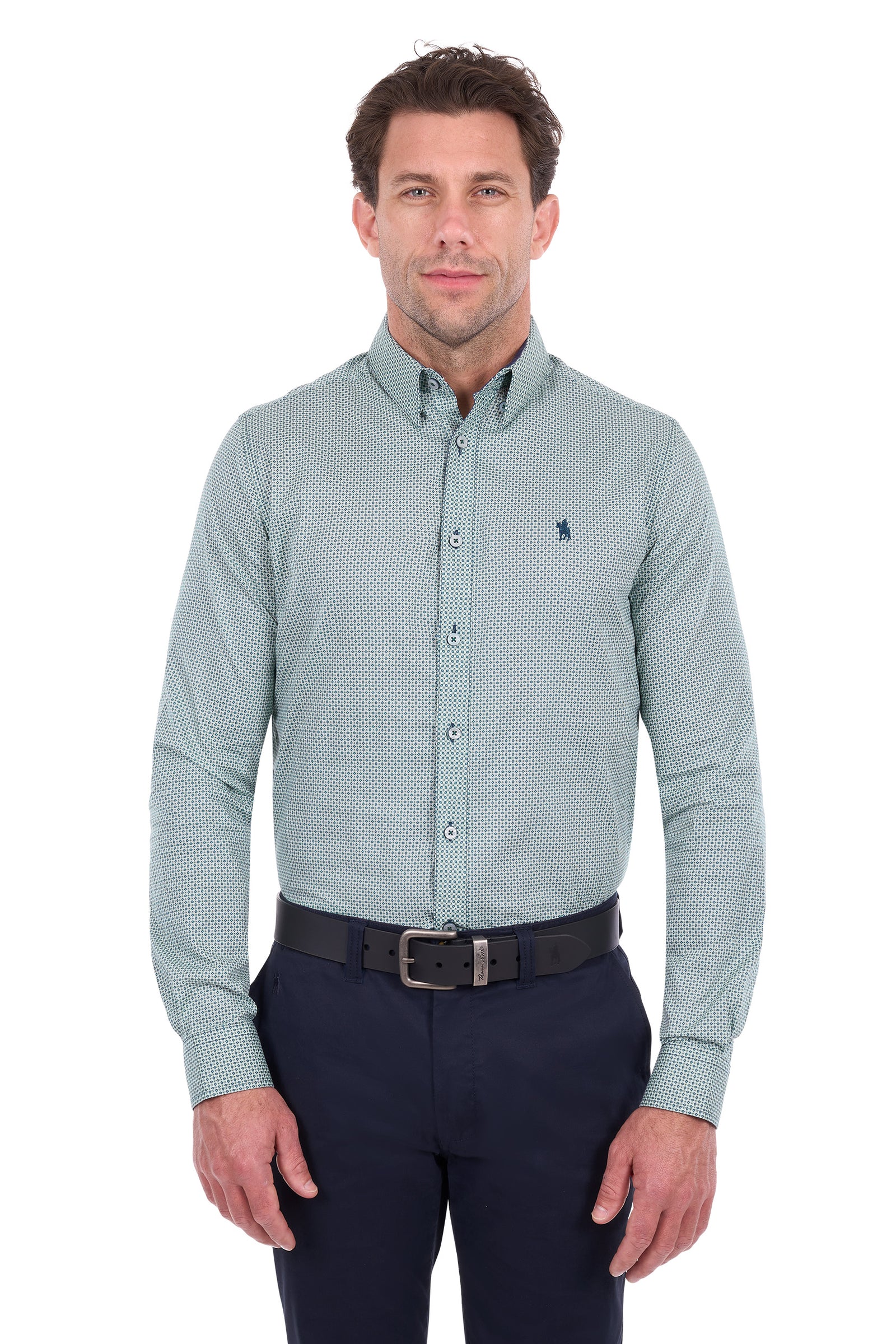 A man wearing a light sage green long-sleeve button-down shirt with a small geometric pattern, paired with navy trousers and a black belt, against a plain white background.