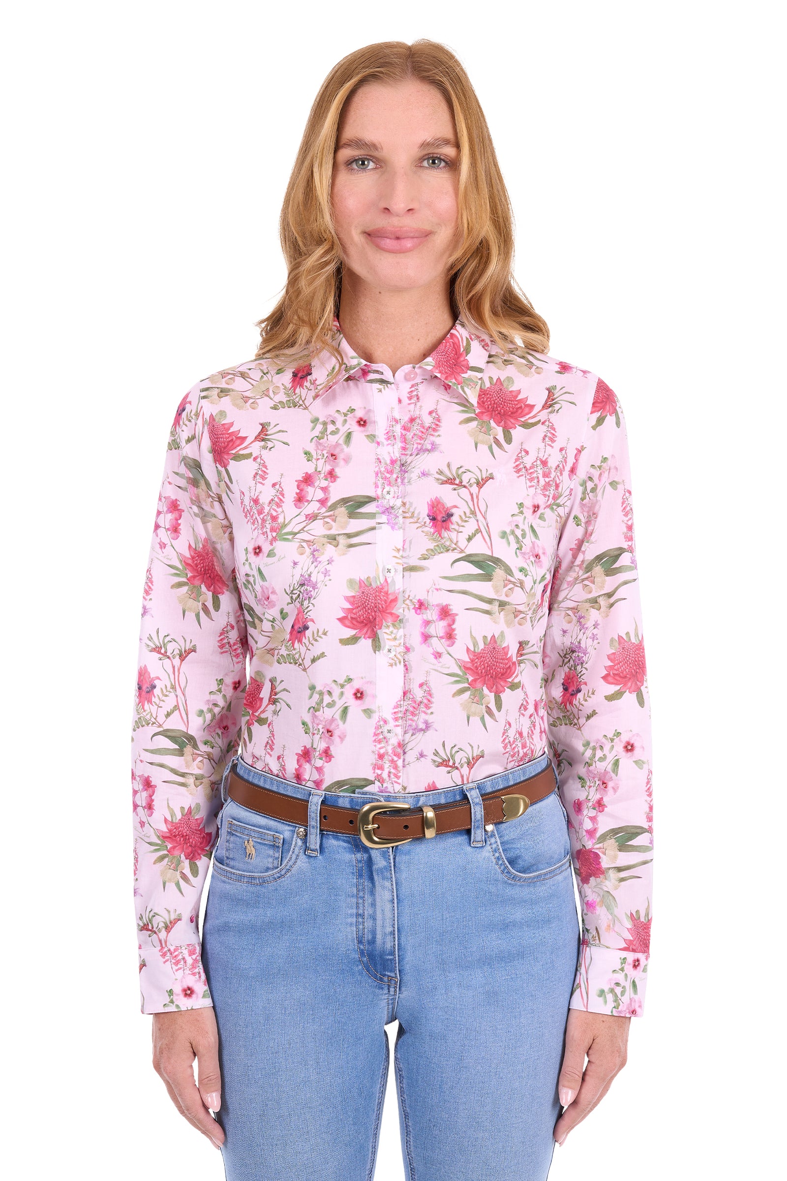 A woman modeling a light pink long-sleeve button-up shirt with a vibrant pink, red, and green floral print, paired with blue jeans and a brown belt, against a plain white background.