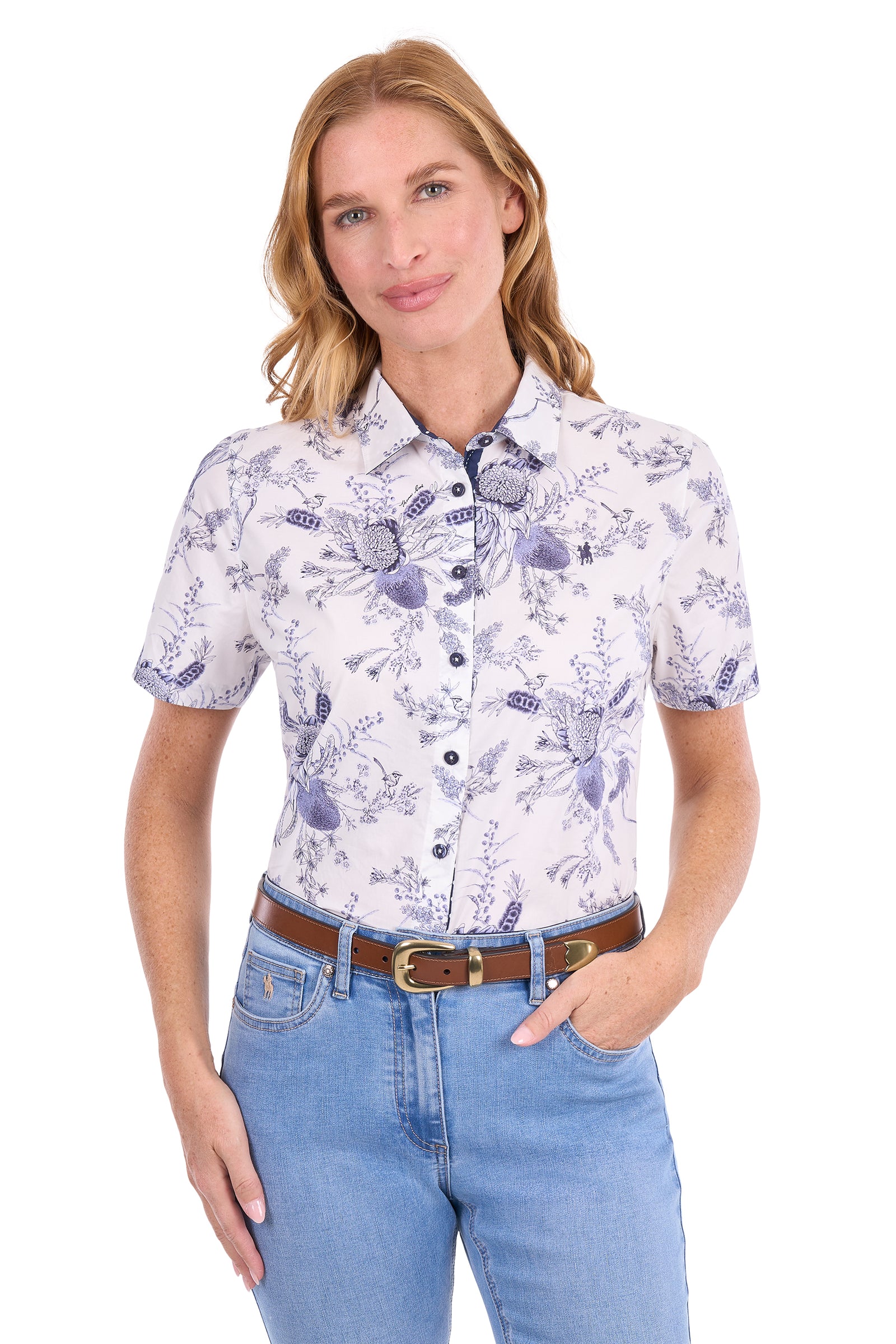A woman modeling a short-sleeve button-up shirt with a white base and a purple floral toile pattern, paired with light blue jeans and a brown belt, against a plain white background.