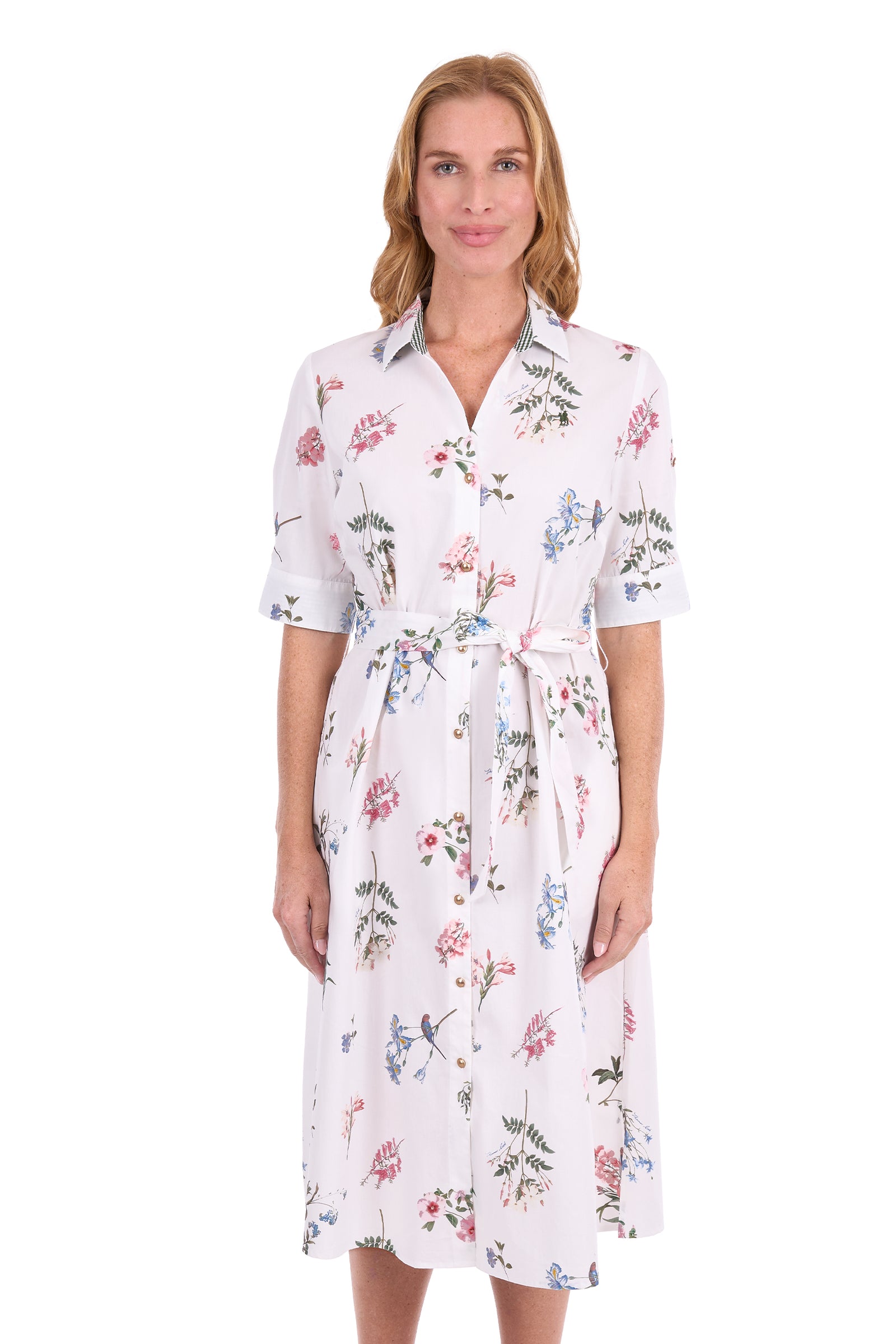 A woman modeling a white short-sleeve collared shirt dress with a matching waist tie and an all-over floral print in pink, blue, and green, set against a plain white background.
