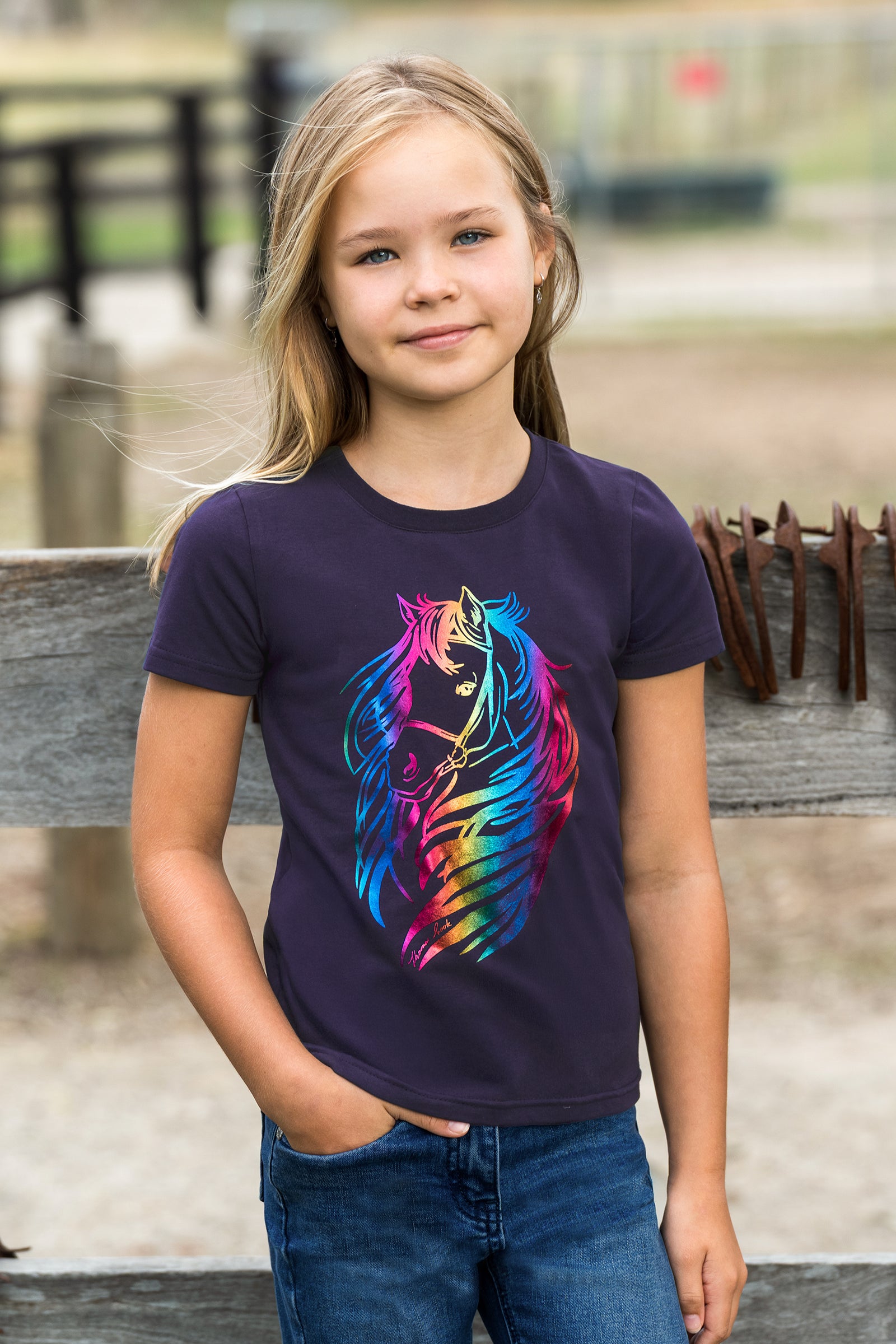 A young girl with blonde hair stands outdoors wearing a dark purple short-sleeve t-shirt with a vibrant, multicolored graphic of a horse head on the front, paired with blue jeans.