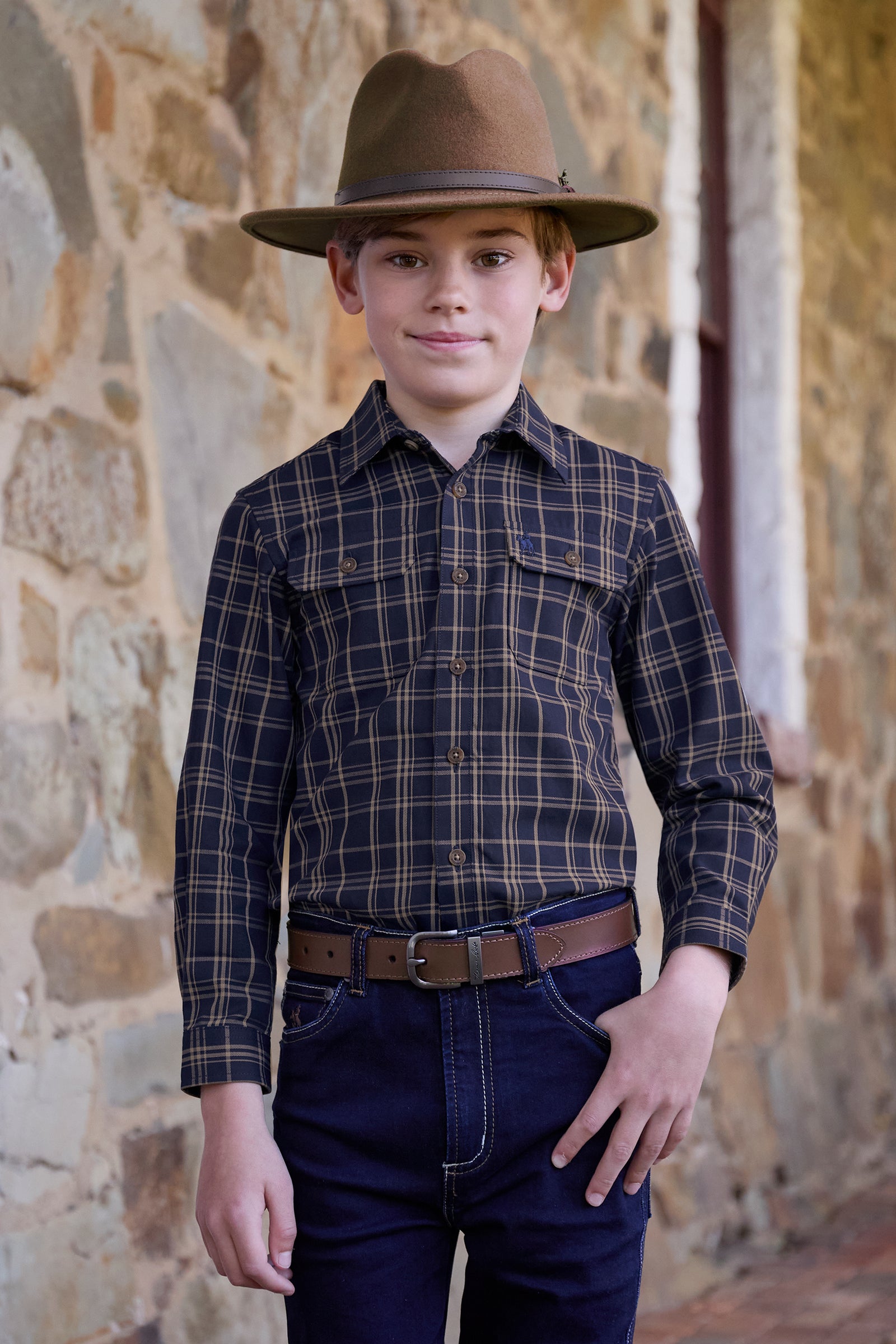 Young boy wearing a plaid shirt, hat, and jeans standing against a stone wall.