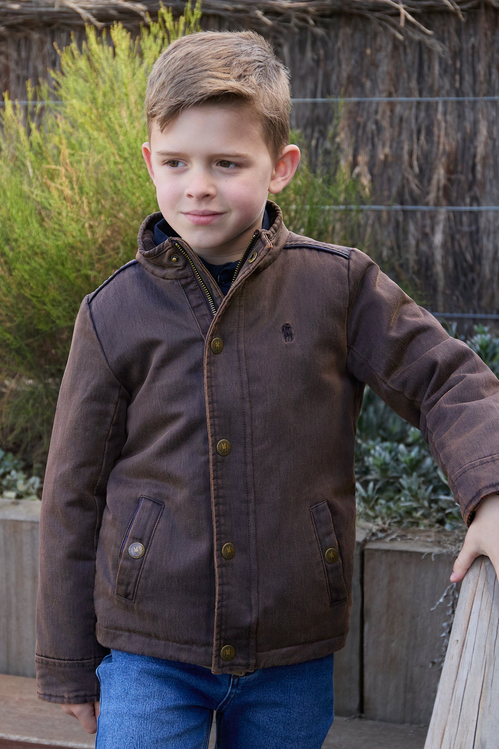 Young boy wearing a brown jacket standing outdoors with plants in the background