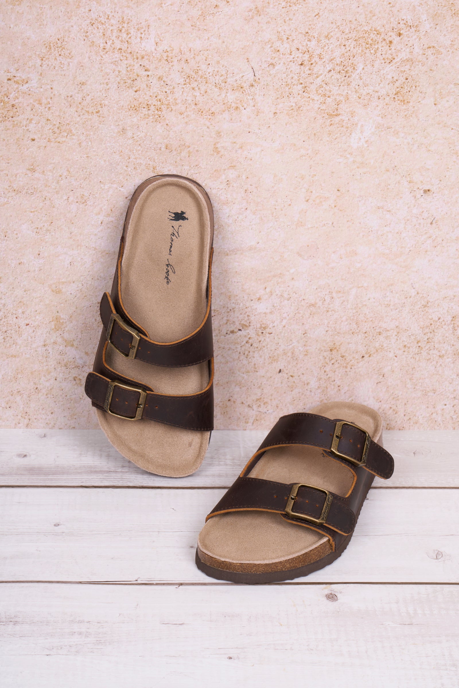 Thomas Cook Women's Trinity Buckle Sandal. Pair of brown sandals with buckles on a light wooden floor and beige wall background