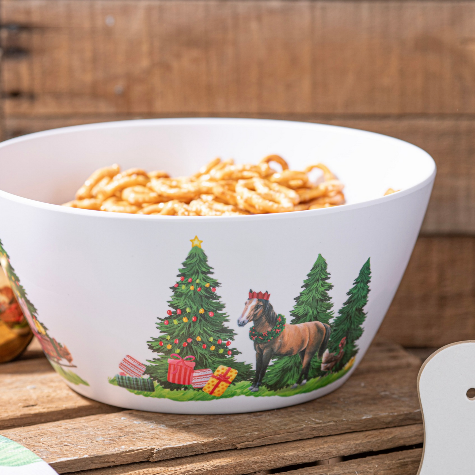 A white ceramic bowl filled with pretzels, decorated with a Christmas tree, horse, and pine tree illustration, set on a rustic wooden surface.