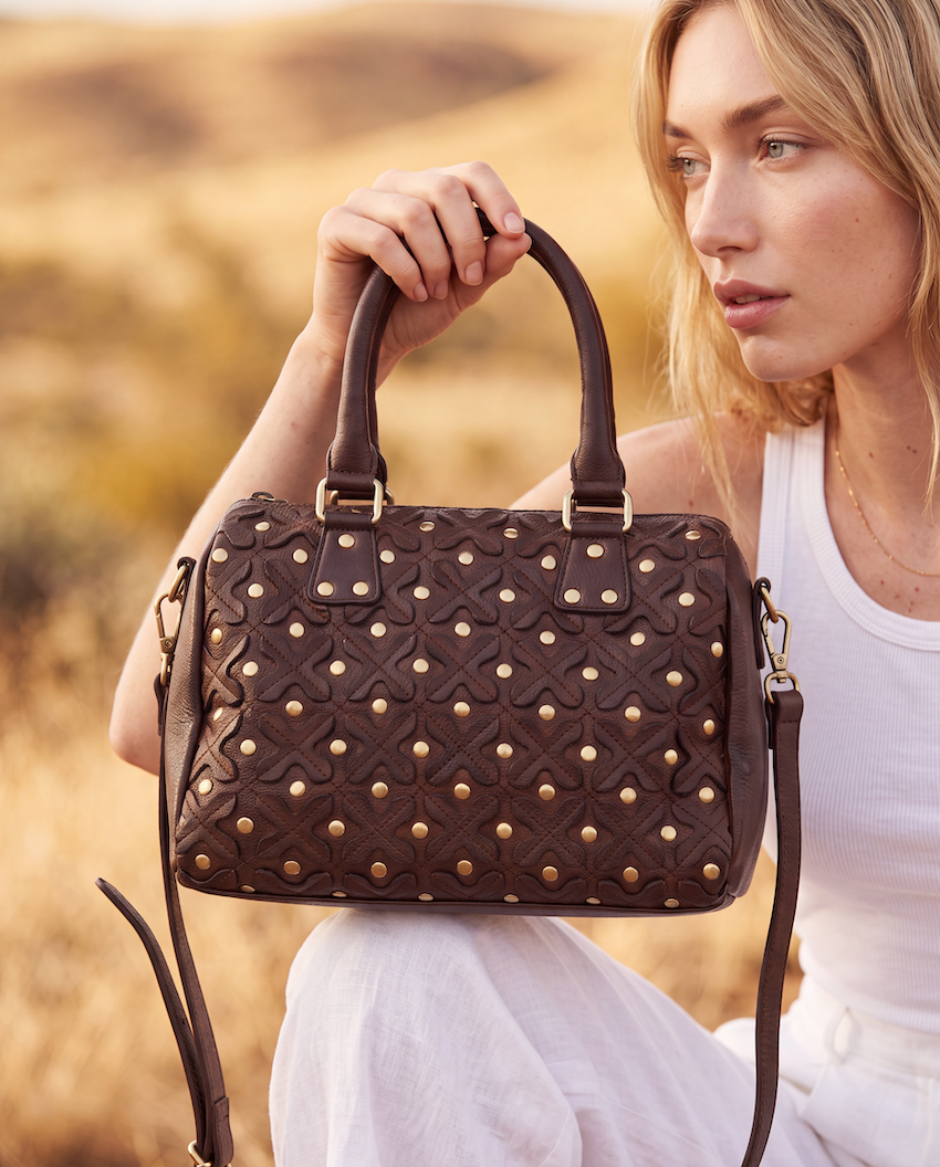 A woman in a white tank top holds a dark brown textured leather handbag with gold-tone studs and top handles, set against a warm, blurred outdoor background.