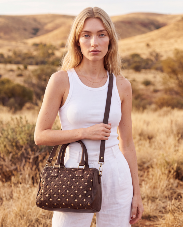 A woman in a white tank top and white pants stands outdoors in a dry grassy landscape, holding a brown quilted shoulder bag with gold-tone studs and a matching brown strap.