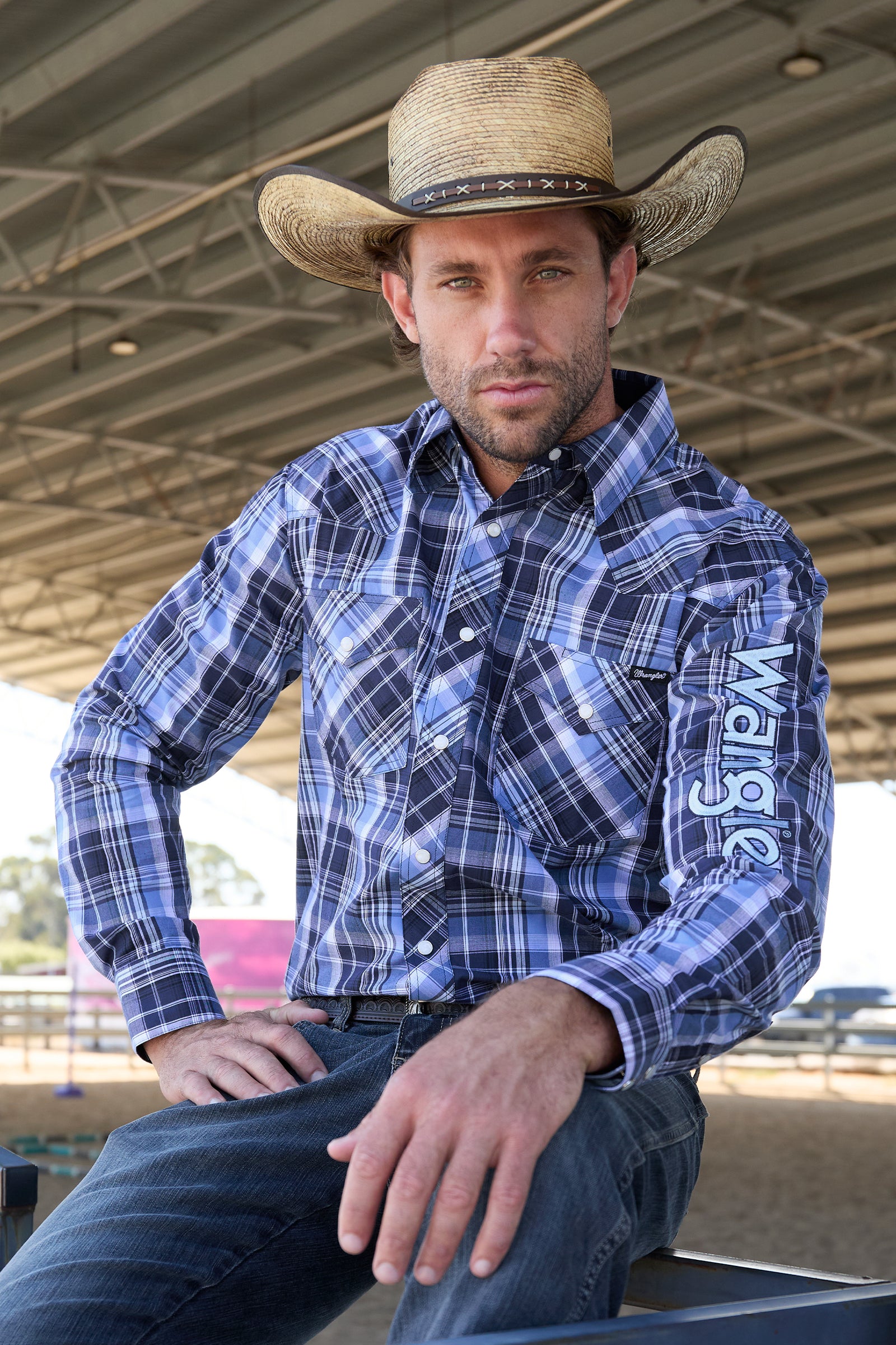 Wrangler Men's Billy Logo L/S Western Shirt. Man wearing a blue plaid shirt with 'Wrangler' branding and a cowboy hat indoors.