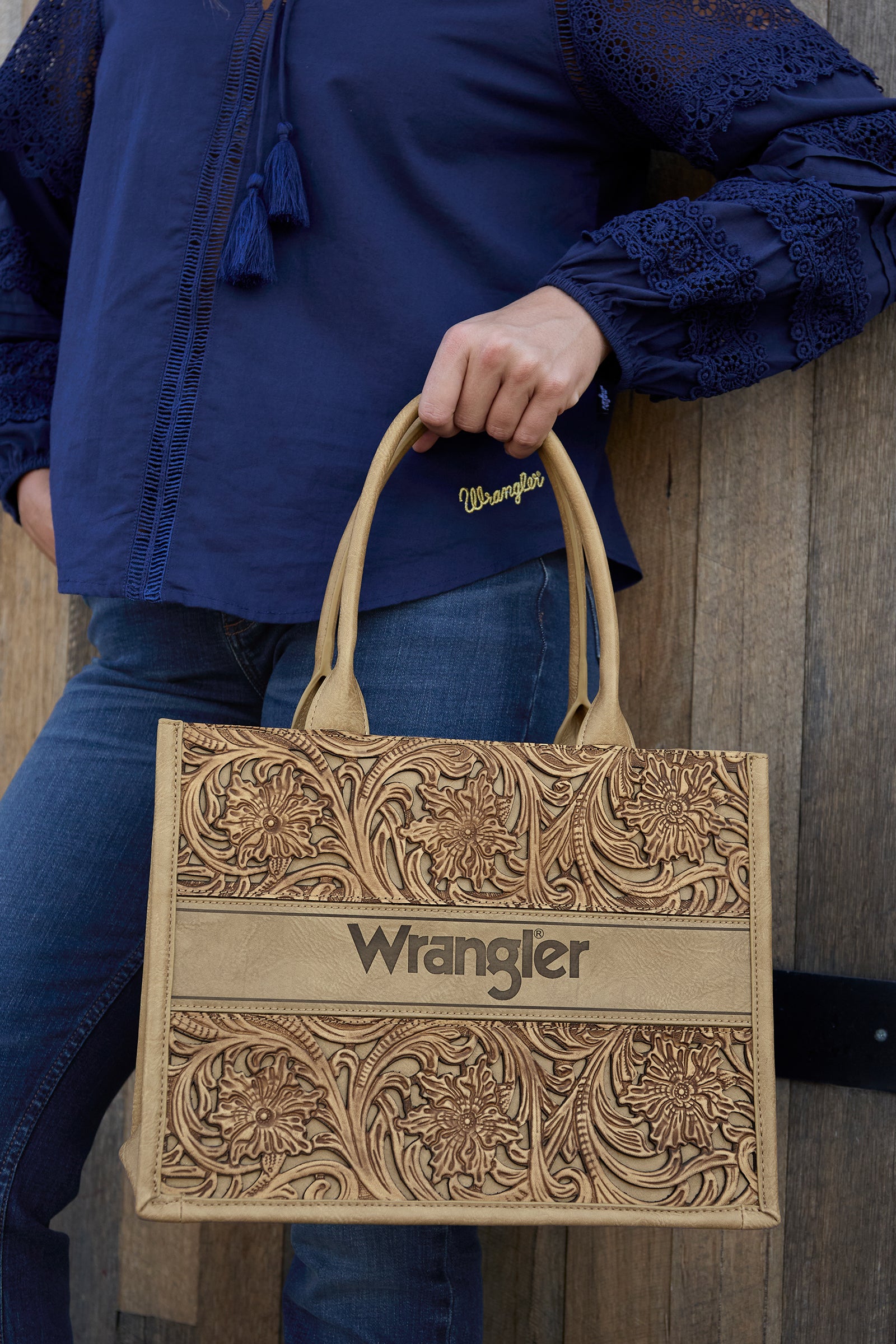 Wrangler Women's Carly Western Bag. Person holding a brown leather bag with 'Wrangler' branding against a wooden background