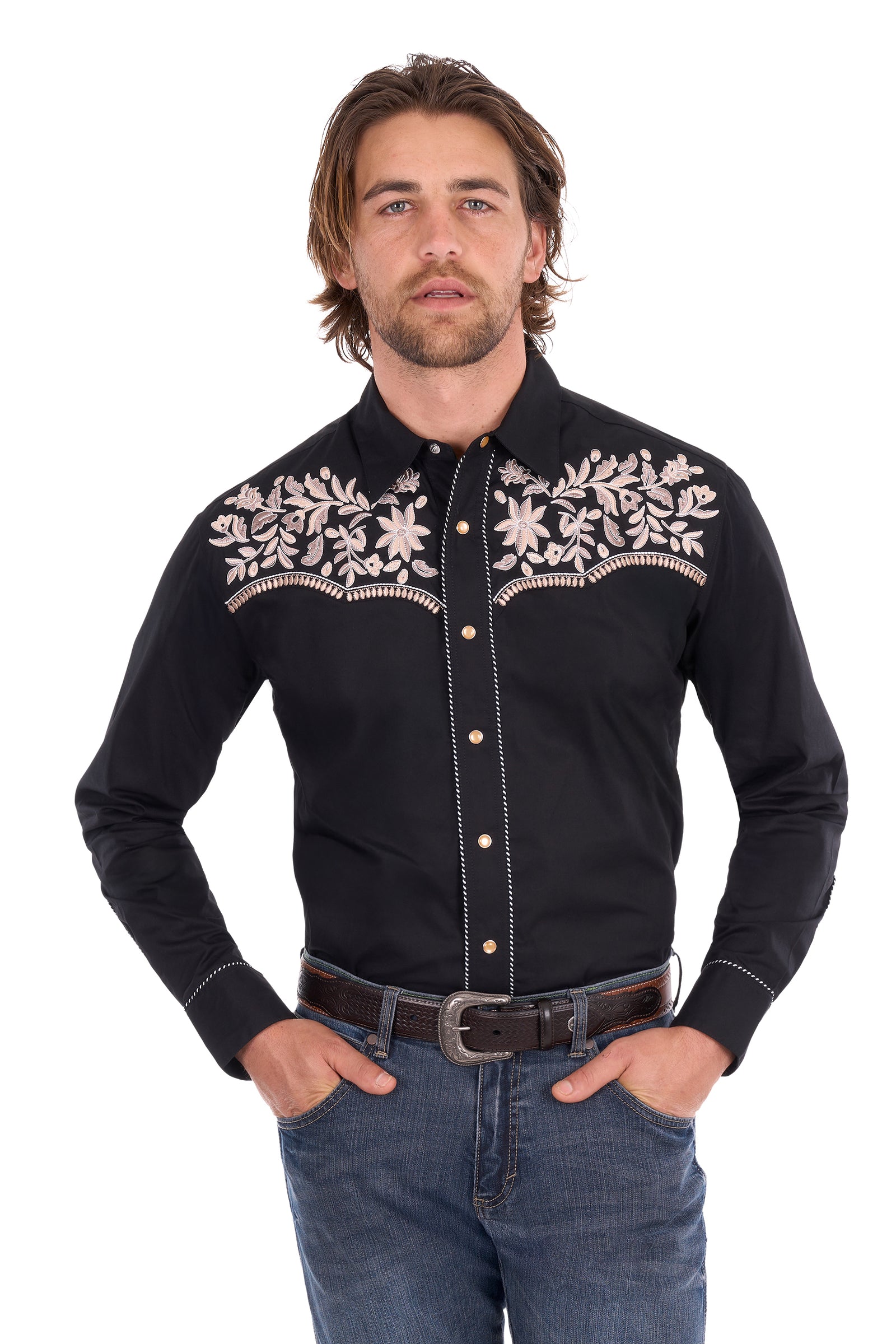 A man wearing a black long-sleeve western shirt with intricate beige floral embroidery across the chest, paired with blue jeans and a brown belt, against a plain white background.