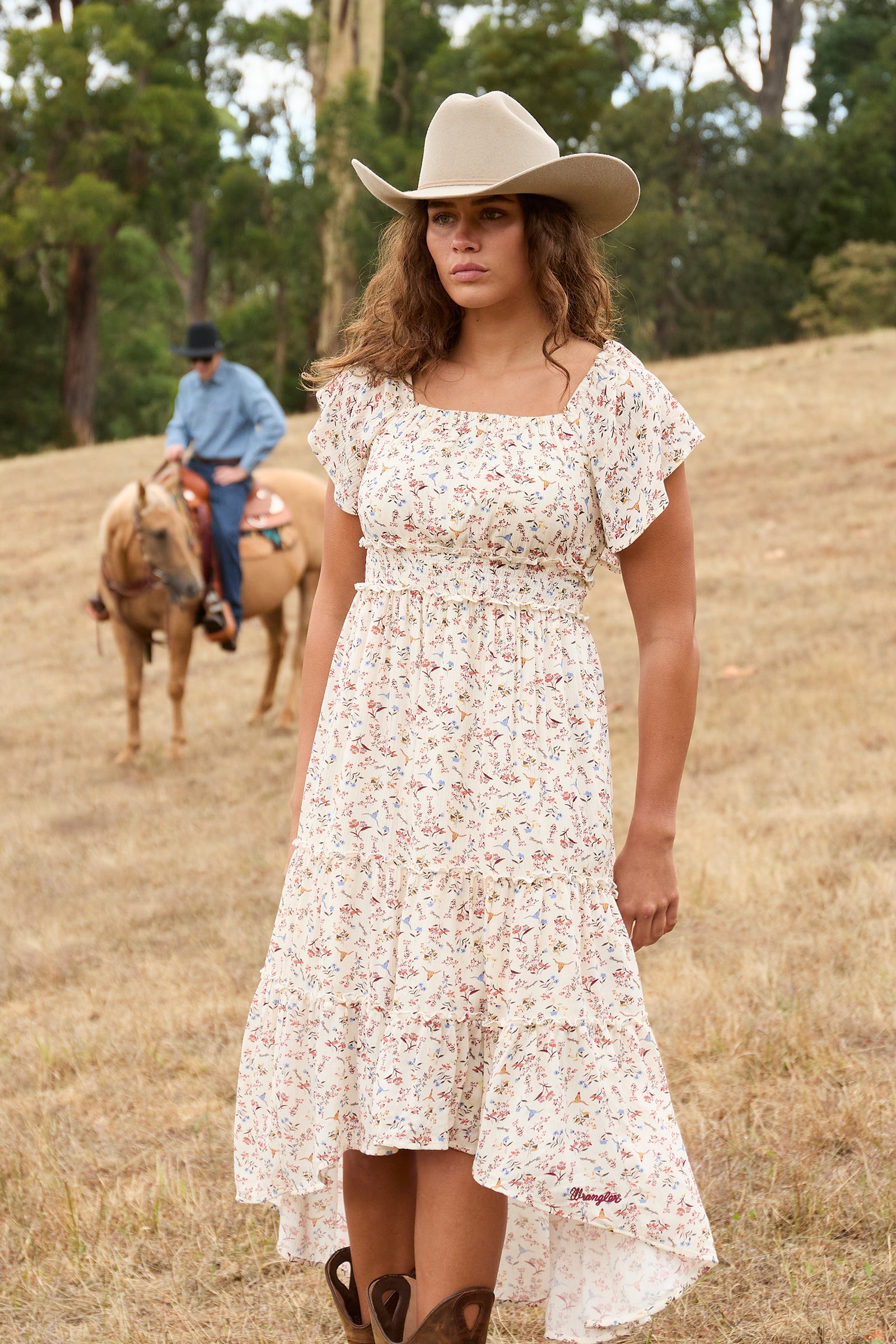 A woman wearing a short-sleeve, high-low floral dress and a beige cowboy hat stands in a dry grass field with trees in the background. A man on a horse is visible in the blurred background.