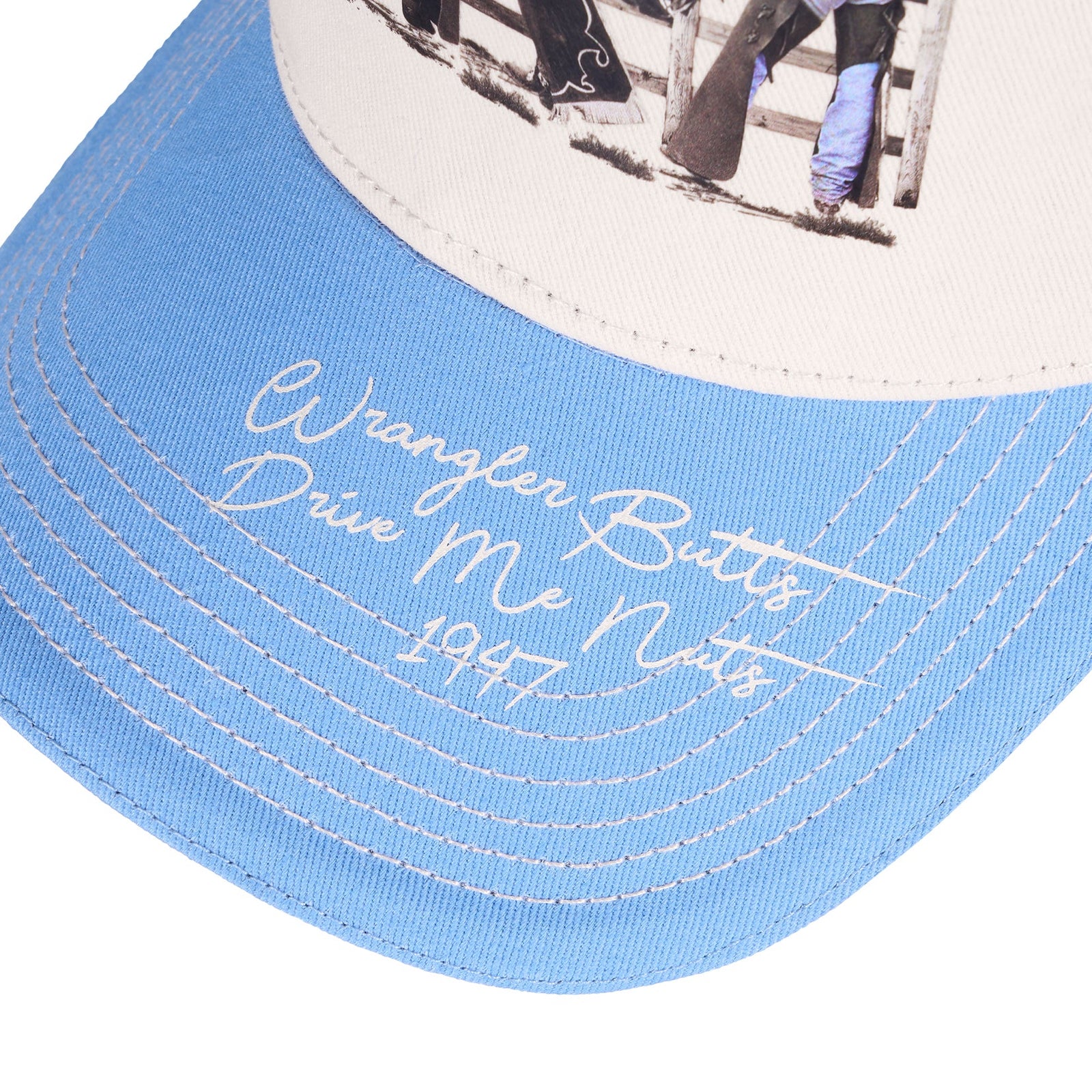 Close-up of a light blue and off-white baseball cap with white embroidered script reading 'Wrangler Butts Drink Me Nots 1947' on the brim.