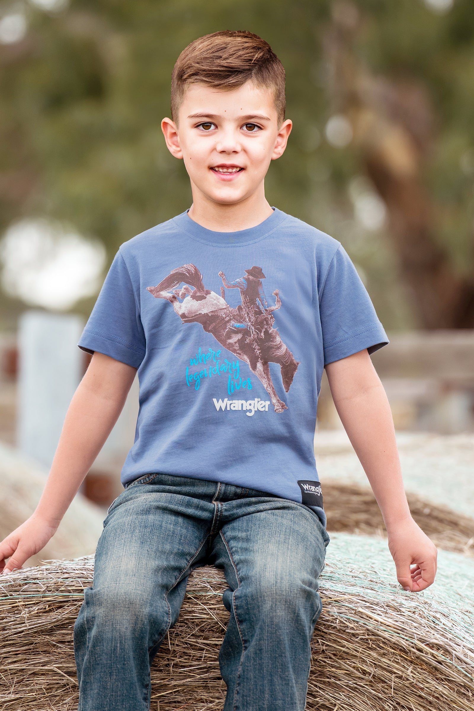 A young boy wearing a blue Wrangler t-shirt with a cowboy riding a bucking bronco graphic, sitting on a hay bale outdoors.