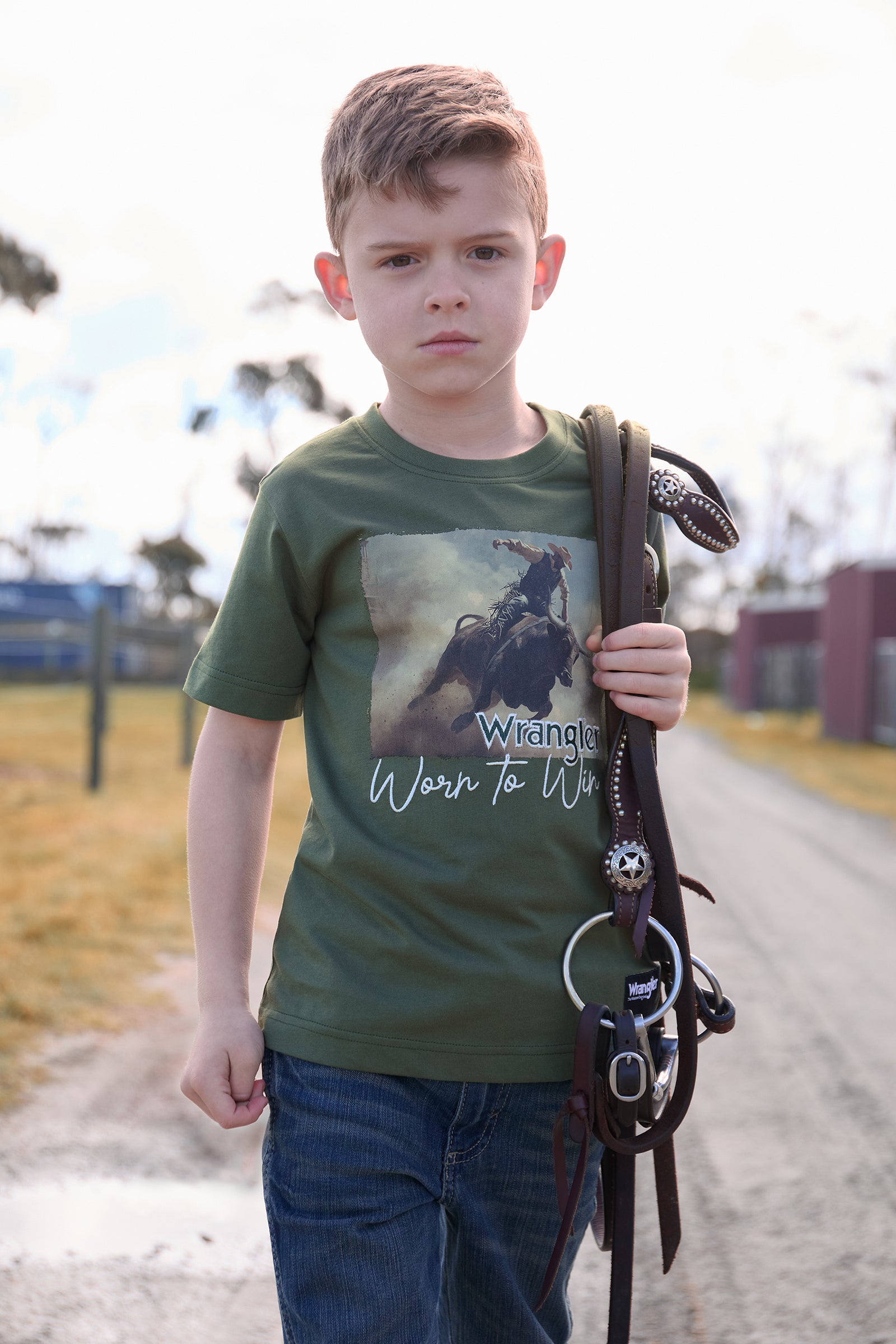 Young boy wearing a green t-shirt with a graphic design, holding a whip on a road.