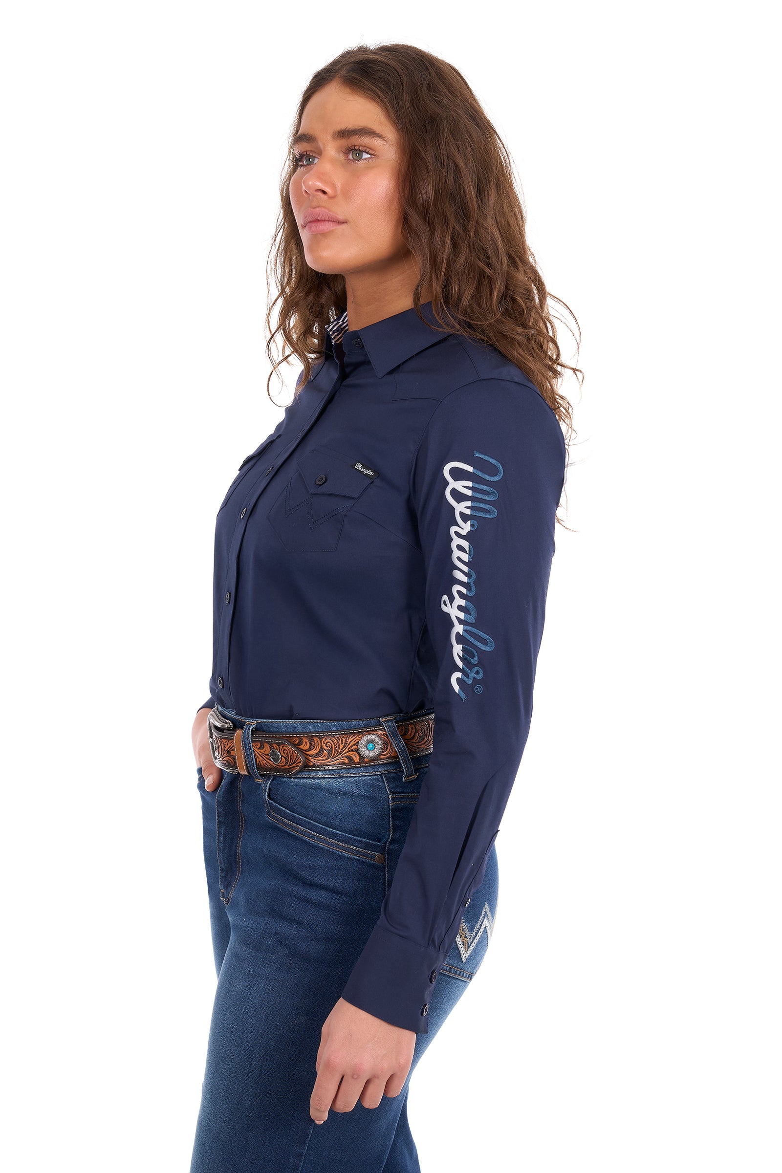 A woman wearing a navy blue long-sleeve western shirt with 'Wrangler' embroidered in white on the sleeve, paired with blue jeans and a brown patterned belt, shown against a plain white background.