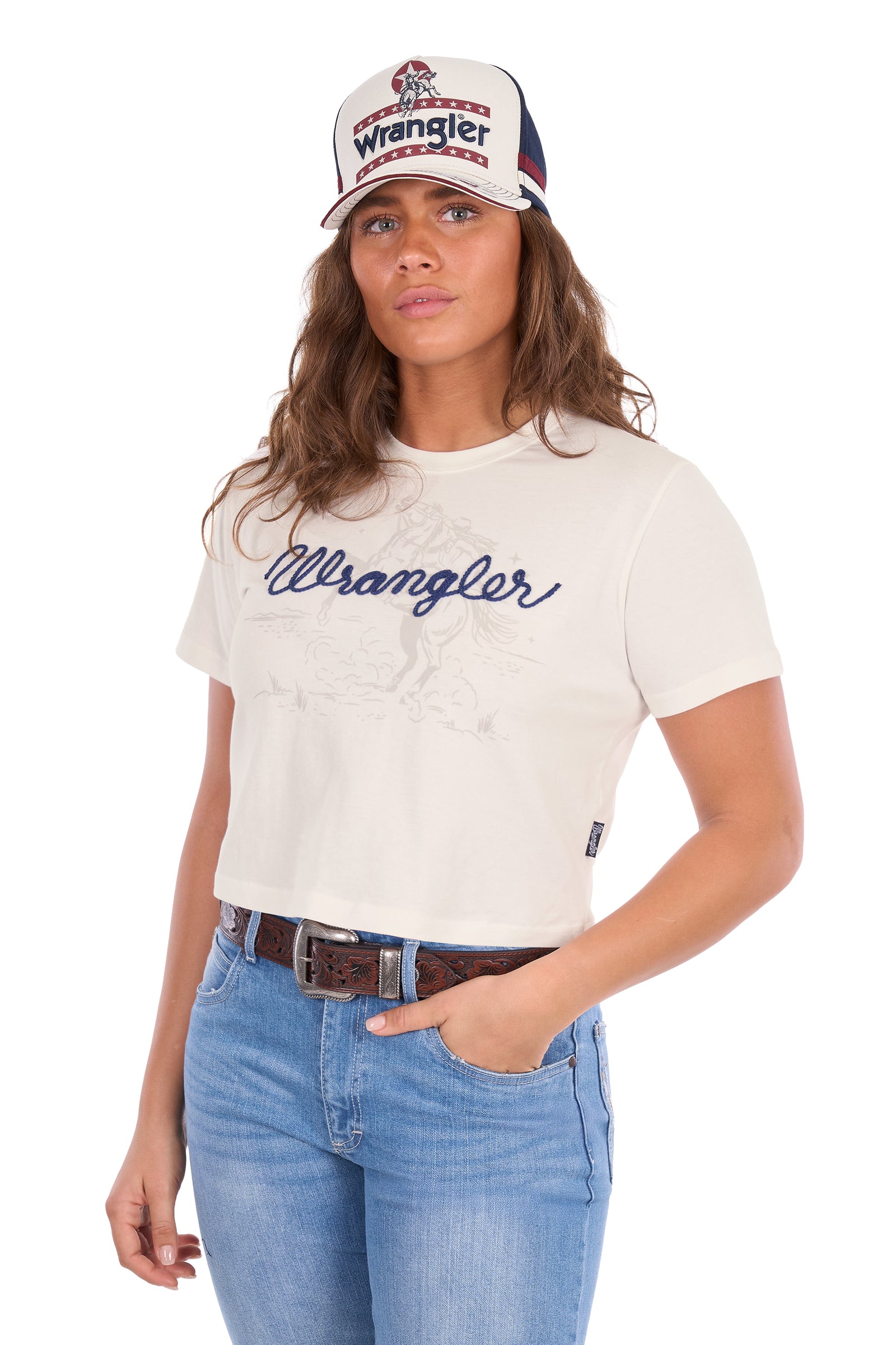 A woman wearing a white Wrangler short-sleeve crop top t-shirt with a navy blue script logo, paired with blue jeans and a white Wrangler trucker hat, against a plain white background.