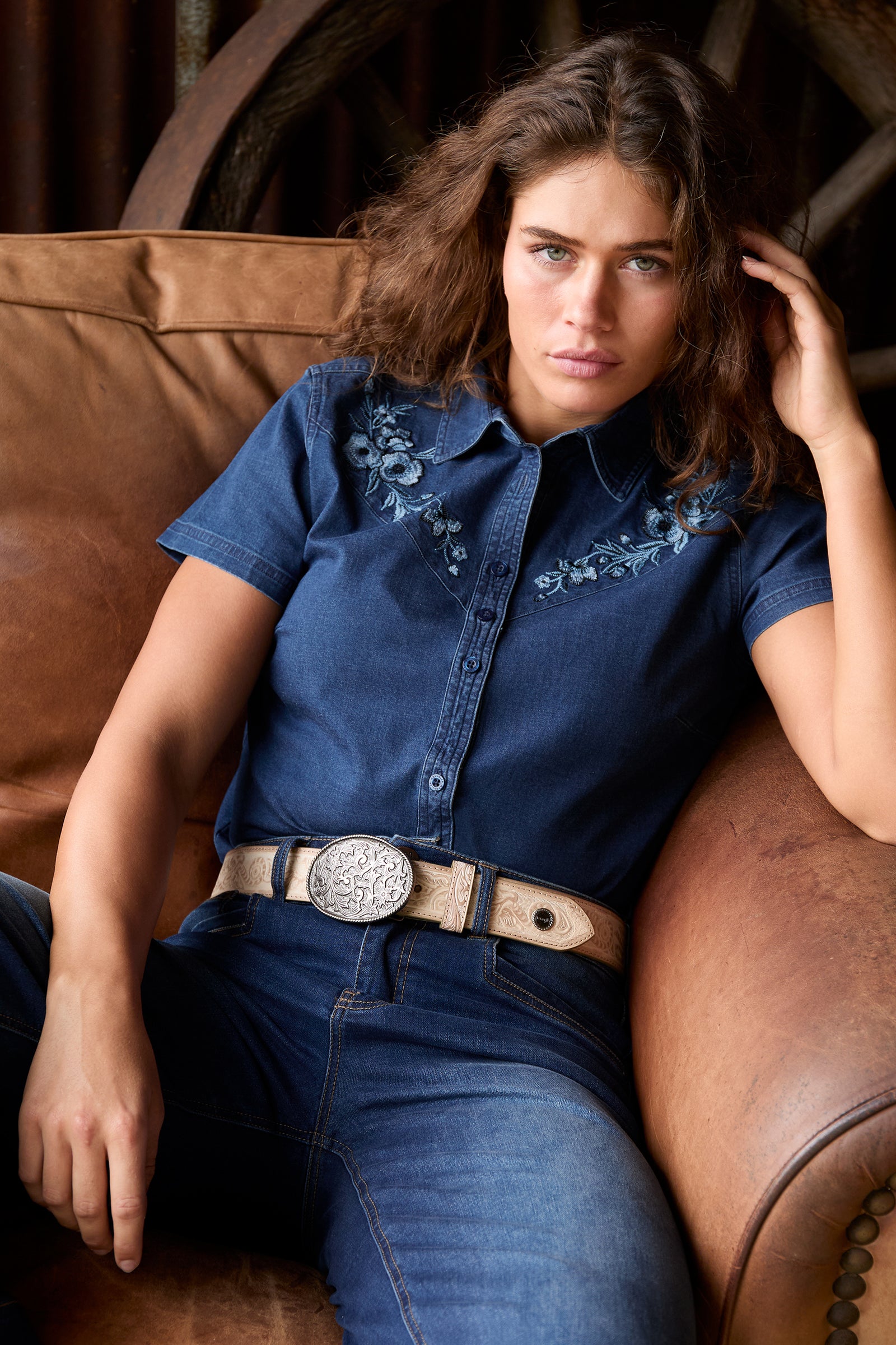 A woman sits on a brown leather couch wearing a blue denim short-sleeve button-up shirt with floral embroidery on the chest, paired with blue jeans and a tan leather belt with a large silver concho buckle.
