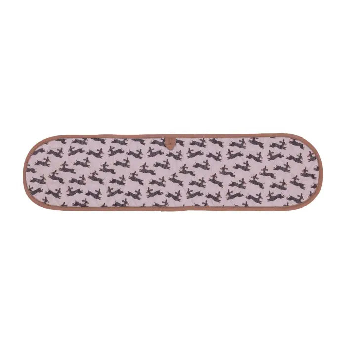 A rectangular quilted blanket with rounded corners, featuring a light pink background with a repeating pattern of black silhouettes of dogs, finished with a brown leather trim.