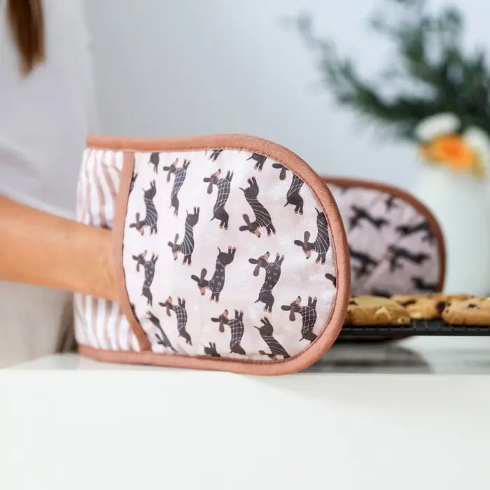Pair of oven mitts with a dog pattern held by a person in a kitchen setting.