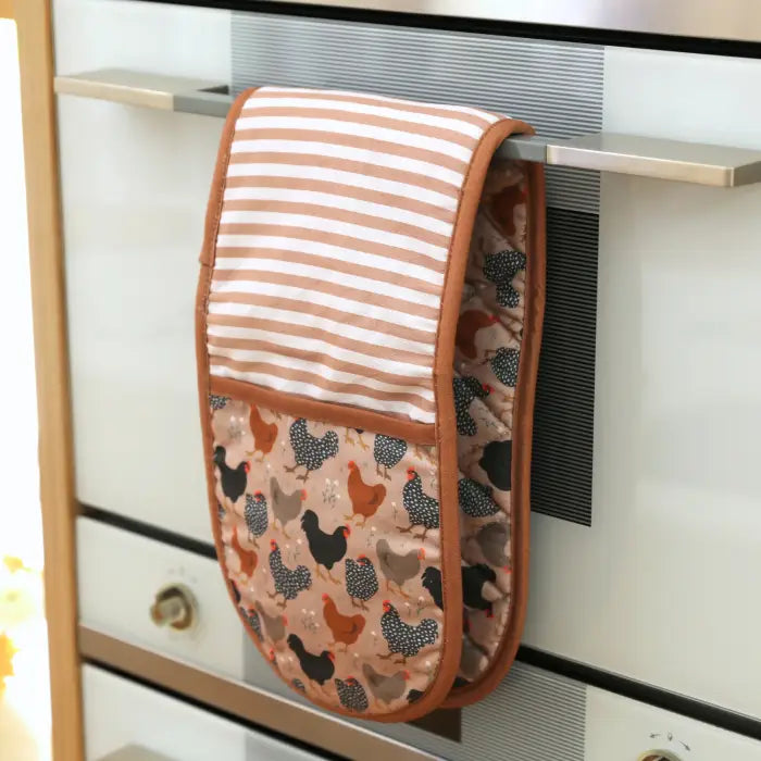 Double oven mitt with chicken pattern hanging on a kitchen cabinet.