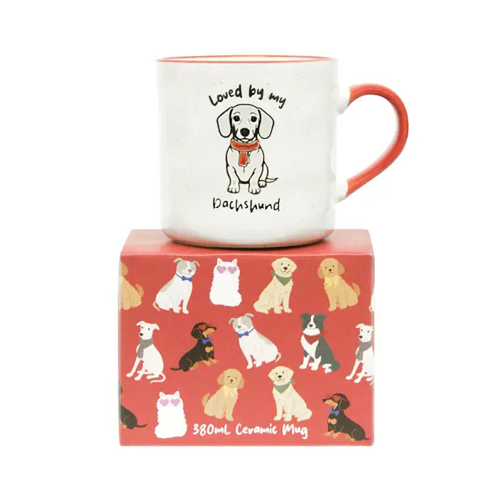 A white ceramic mug with an orange-red handle and interior, printed with a dachshund illustration and the words 'Loved by my Dachshund.' The mug is displayed on top of a red patterned gift box featuring dog illustrations.