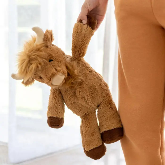 A plush Highland cow stuffed animal being held by a person wearing a tan coat in a bright indoor setting.