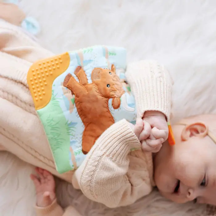 A baby wearing a cream knit outfit lies on a soft white surface, holding a colorful fabric teething toy with a brown plush animal and textured yellow panel.