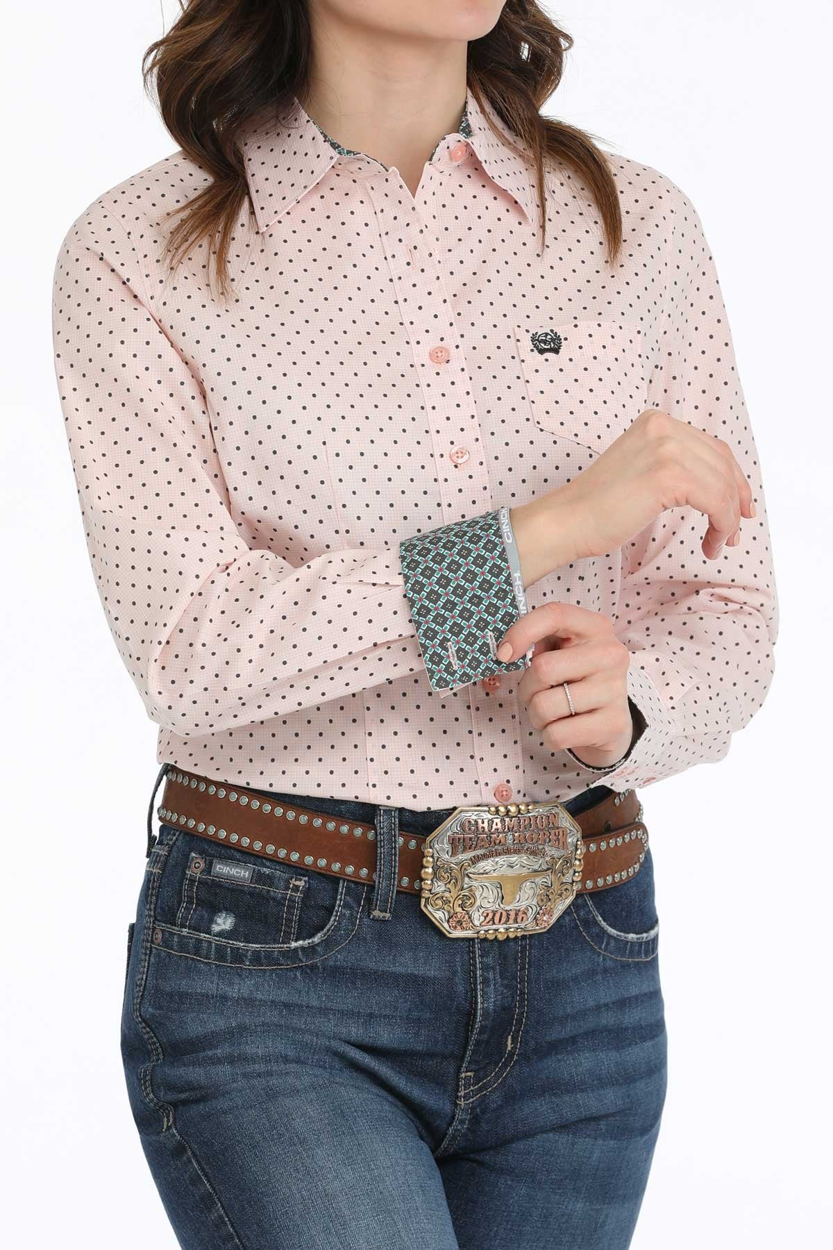 Cinch Women's Geometric Print L/S Western Shirt. Woman wearing a pink patterned shirt and blue jeans with a belt and large buckle.