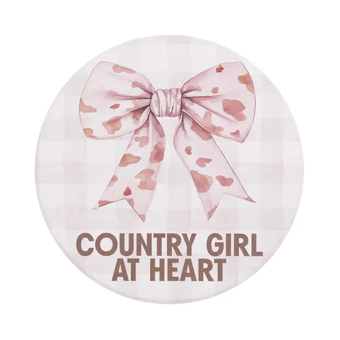 A round decorative sign or plaque featuring a watercolor-style pink bow on a pale pink gingham background, with the text 'COUNTRY GIRL AT HEART' in brown at the bottom.